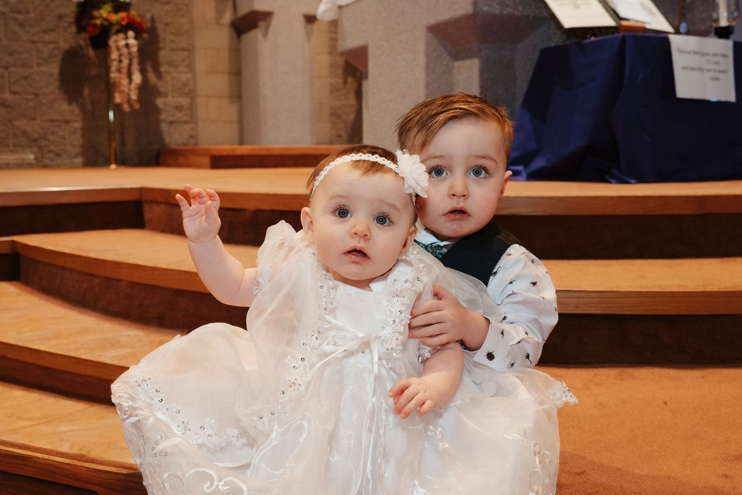 Two young siblings in christening attire sitting together on church altar steps.