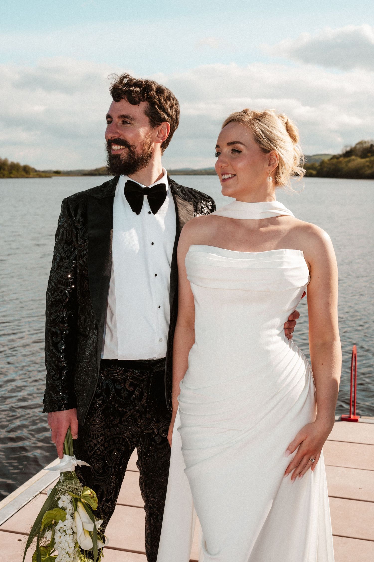 Bride in sleek halter neck gown and bearded groom in black sequin tuxedo pose on lakeside dock.