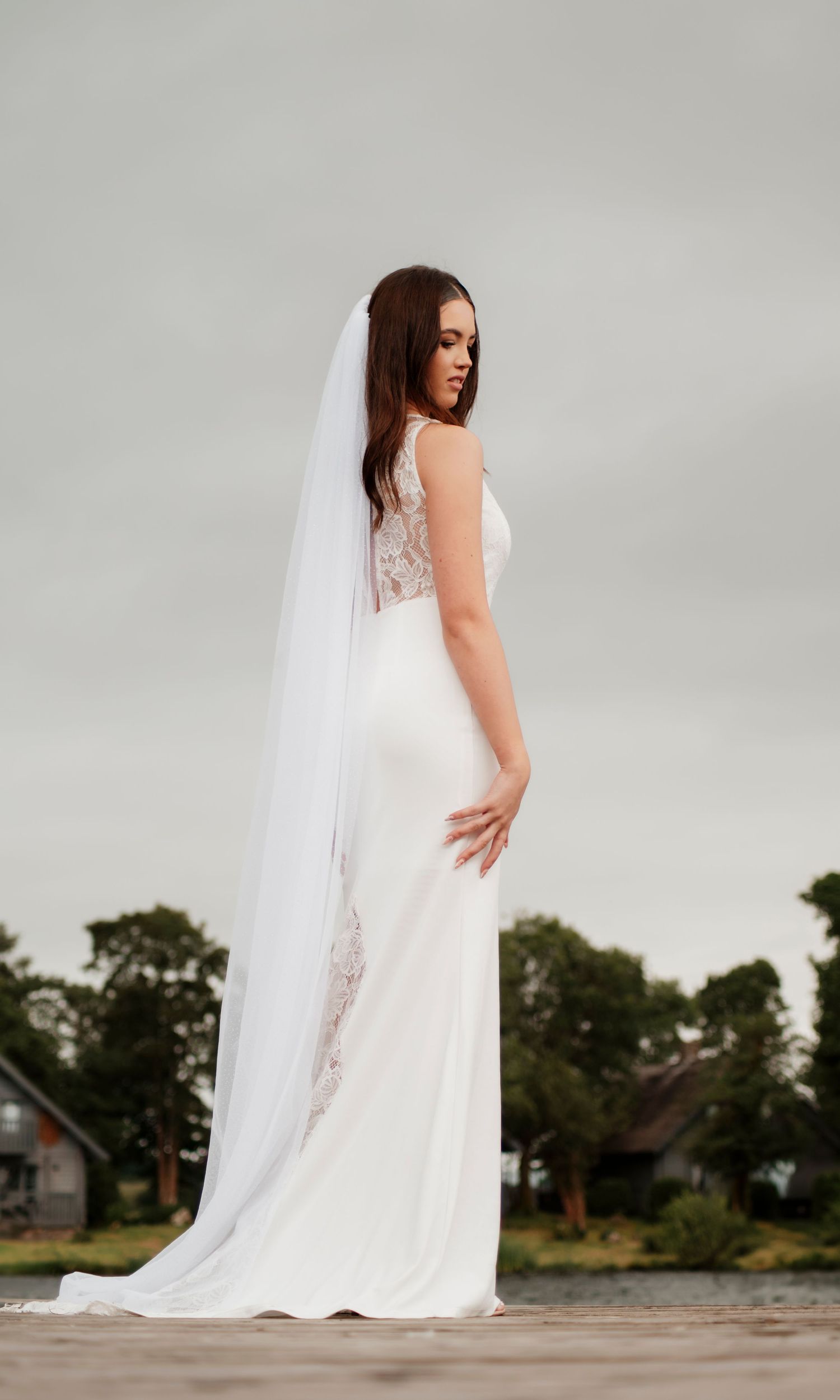 Bride in elegant backless white wedding gown with long cathedral veil stands outdoors on overcast day.