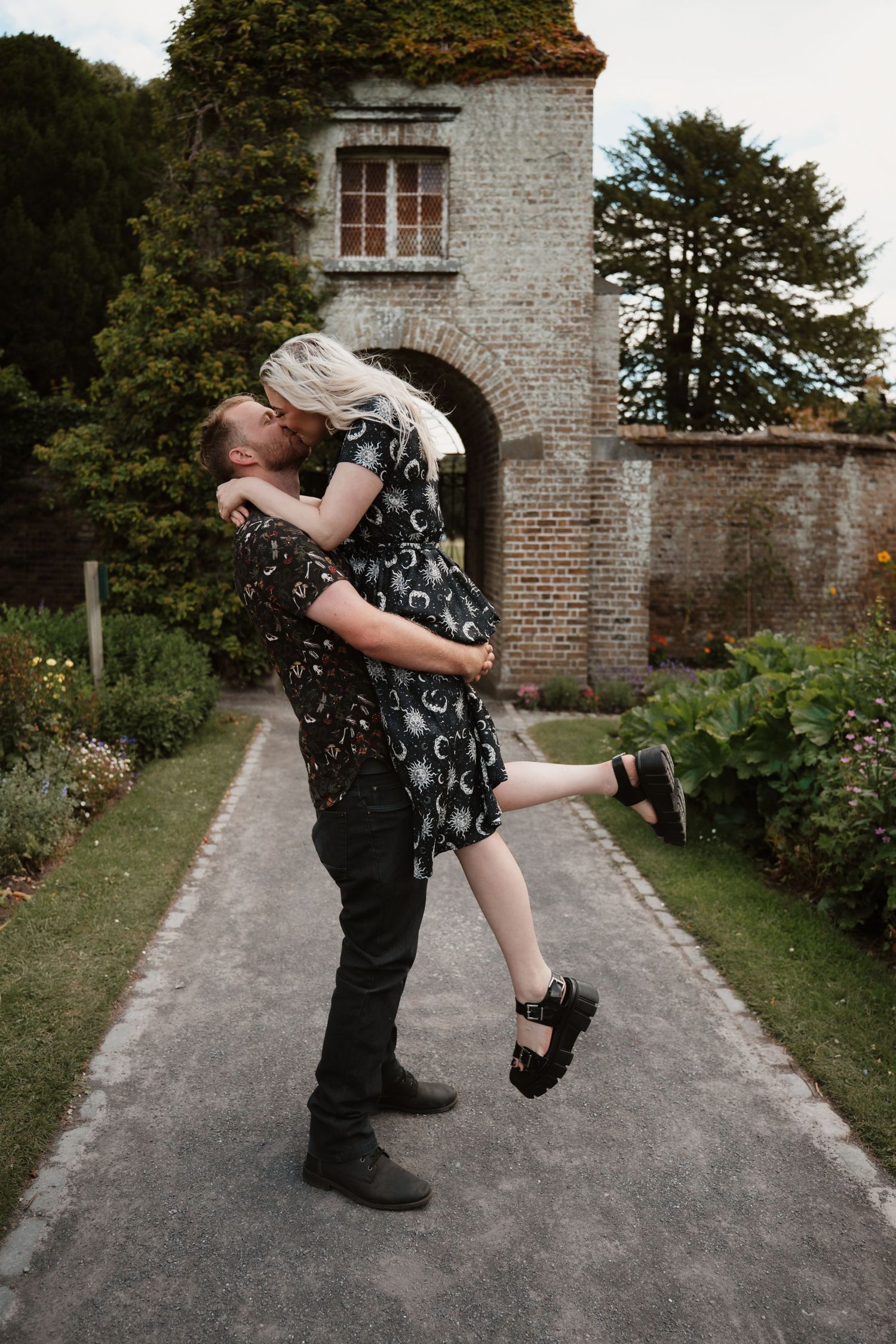 A couple sharing a passionate kiss as the man lifts the woman in a moonprint dress in a lush English garden setting.
