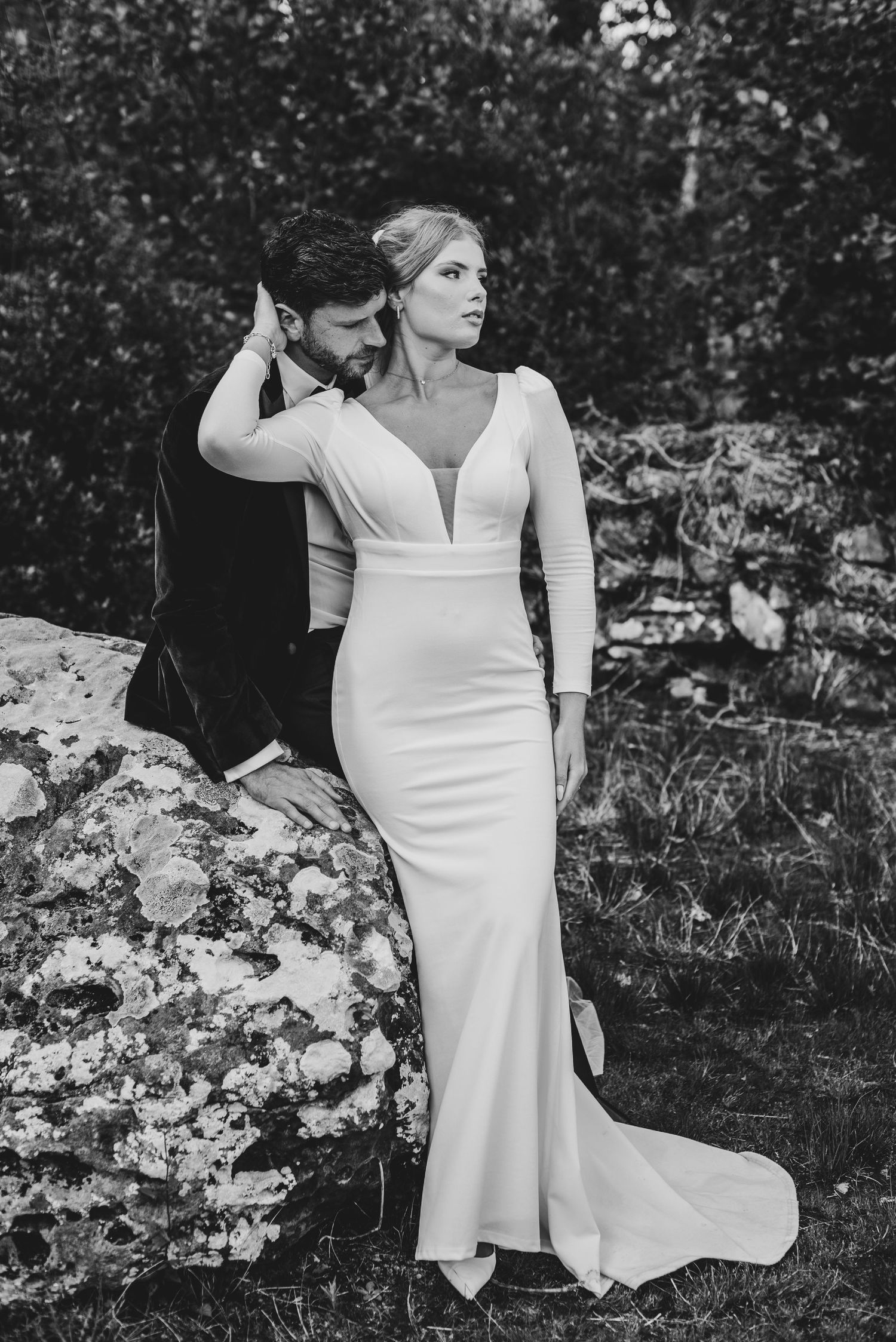 Black and white photo of elegant bride in fitted gown with groom in nature.