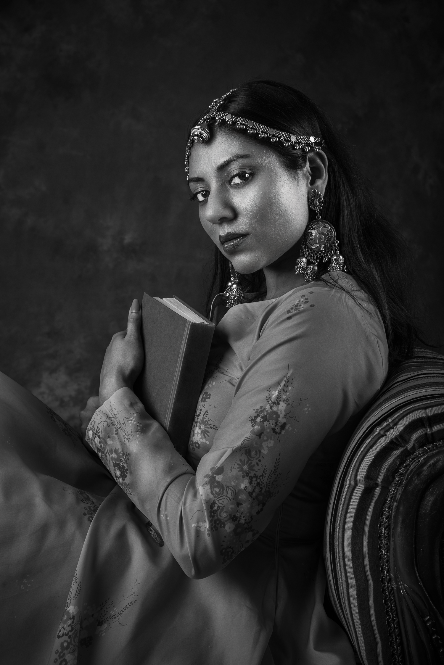 Black and white portrait of a woman in traditional Indian attire holding a book, wearing ornate jewelry and headpiece.