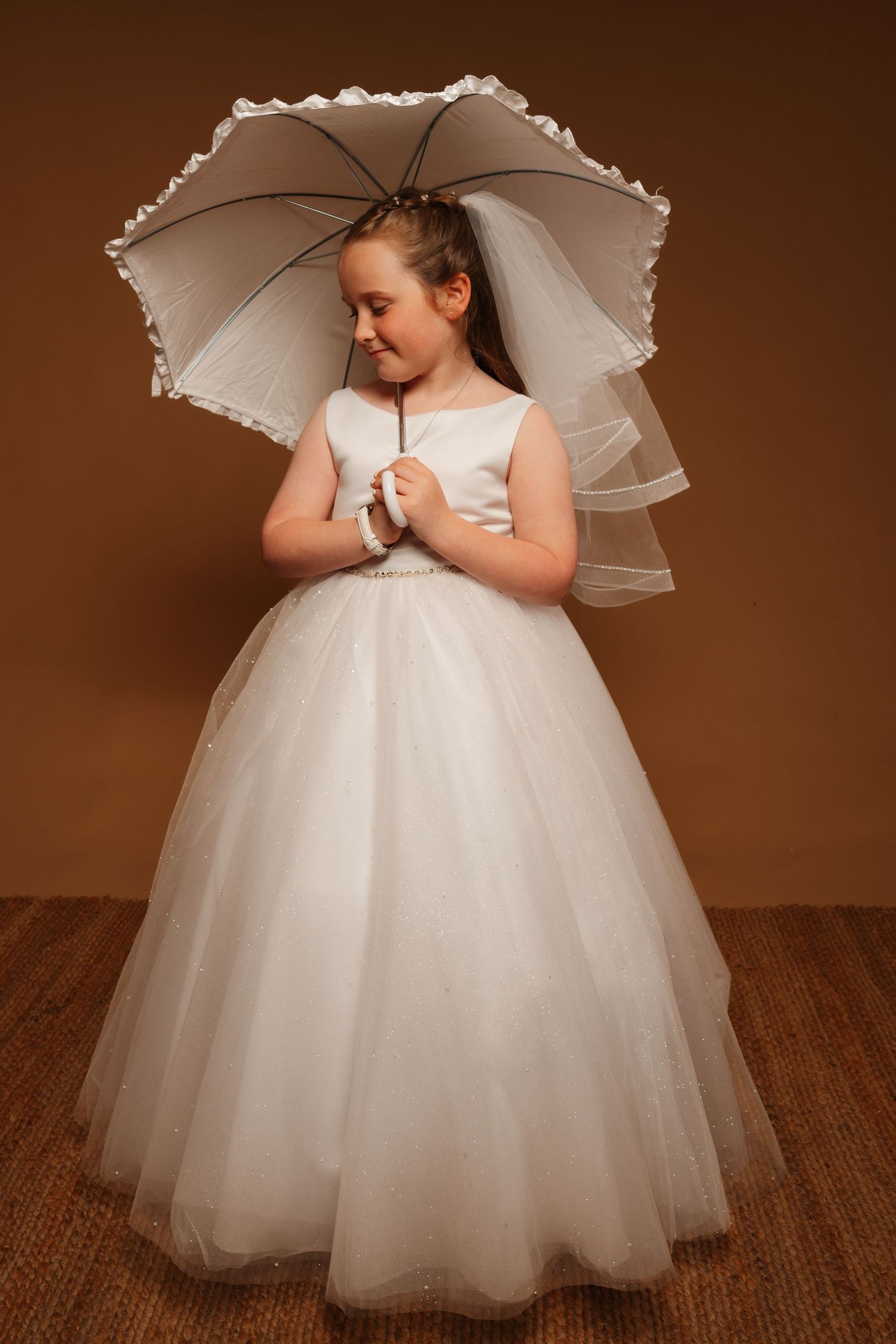 Young girl in white communion dress and veil holds matching parasol umbrella, posing against brown background.