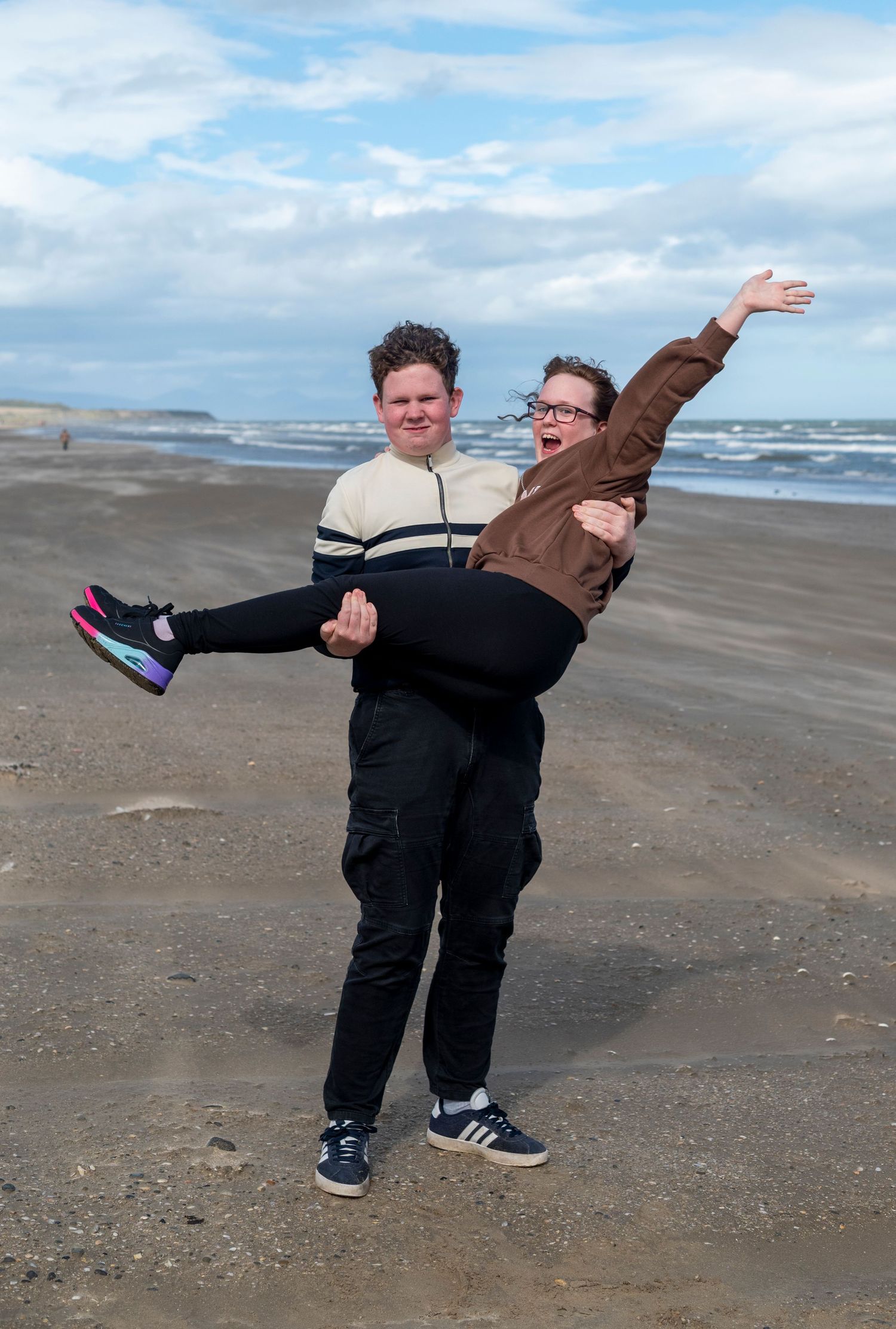Two young people joyfully posing on a sandy beach, one carrying the other in their arms with waves in the background.