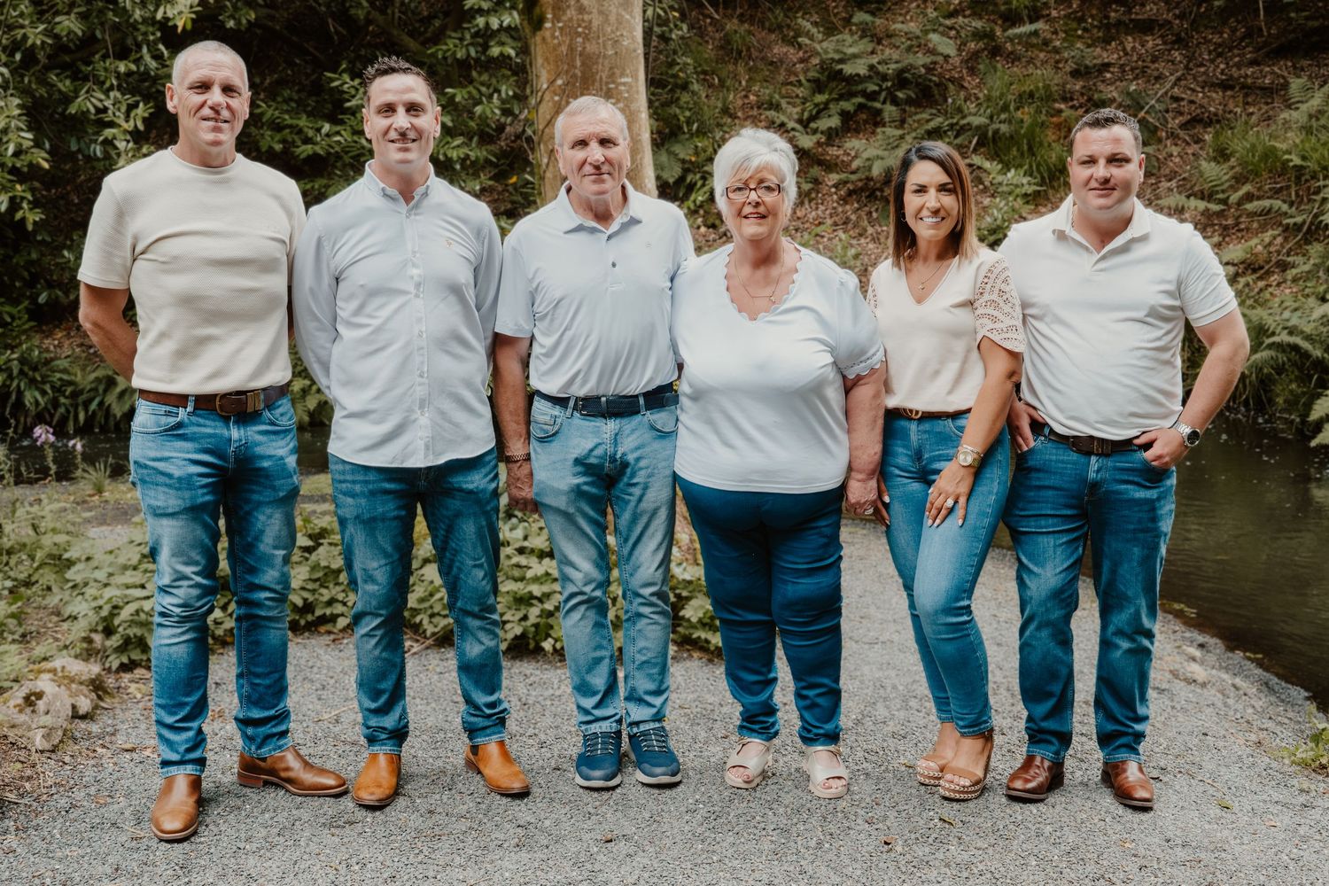 Six adults in matching white polos and jeans pose together outdoors in a lush green setting for a family portrait.