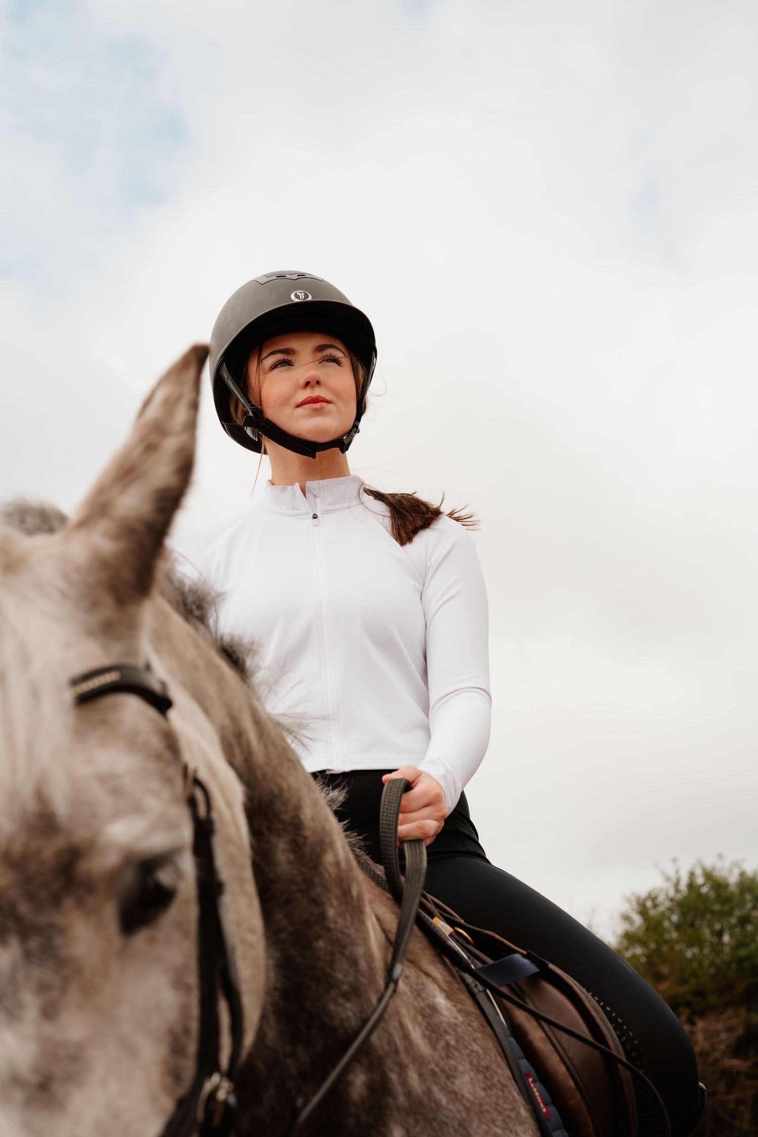 Young woman in black helmet and white top rides a gray horse outdoors under a cloudy sky.