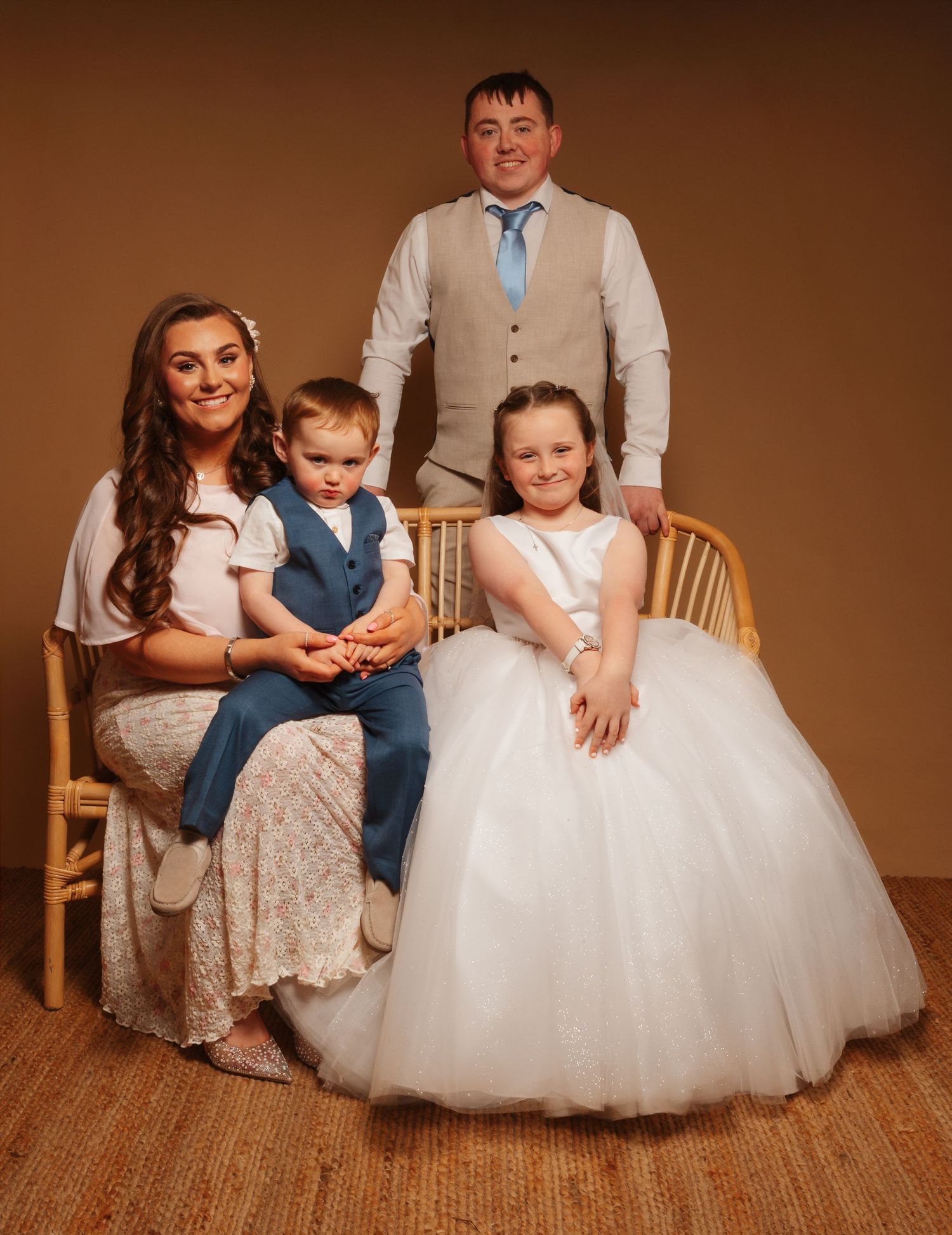 Family portrait with mother, toddler son, daughter in white communion dress, and father posing together for a formal photo.