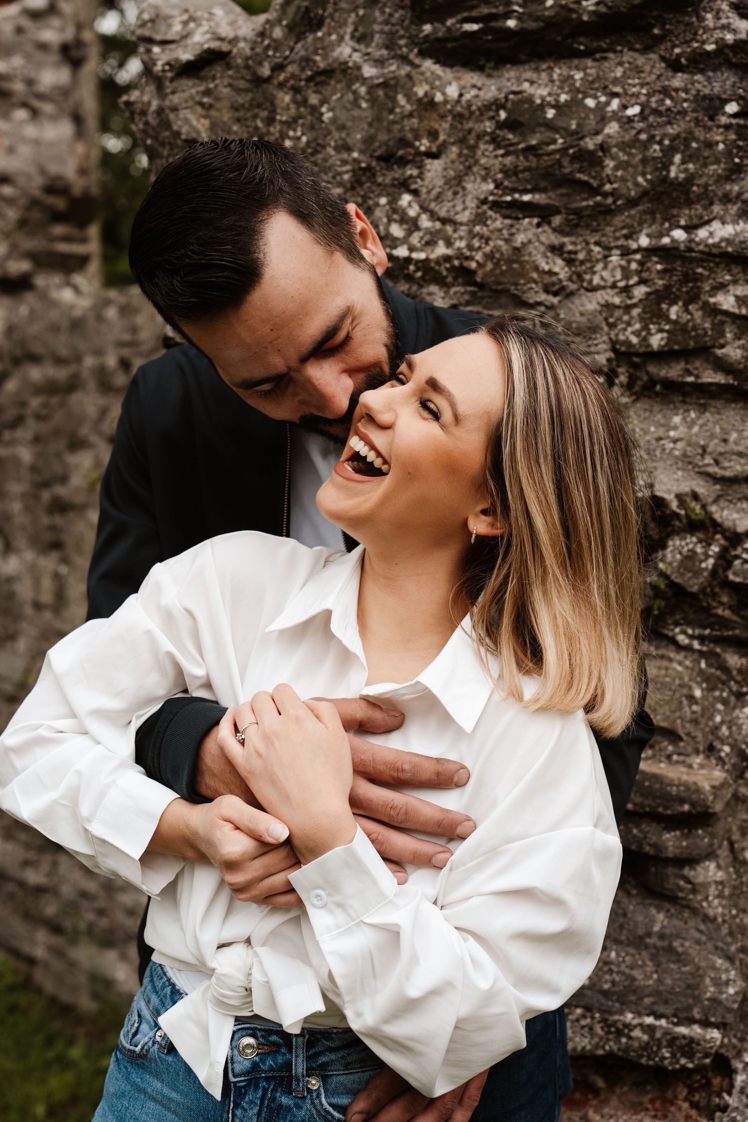A joyful couple laughing and embracing against a rustic stone wall, woman in white shirt and jeans.