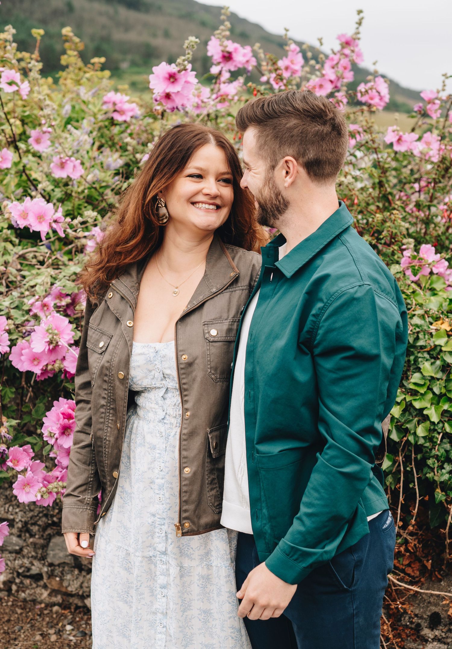 Happy couple standing together in front of pink flowering bushes, woman in floral dress and jacket.