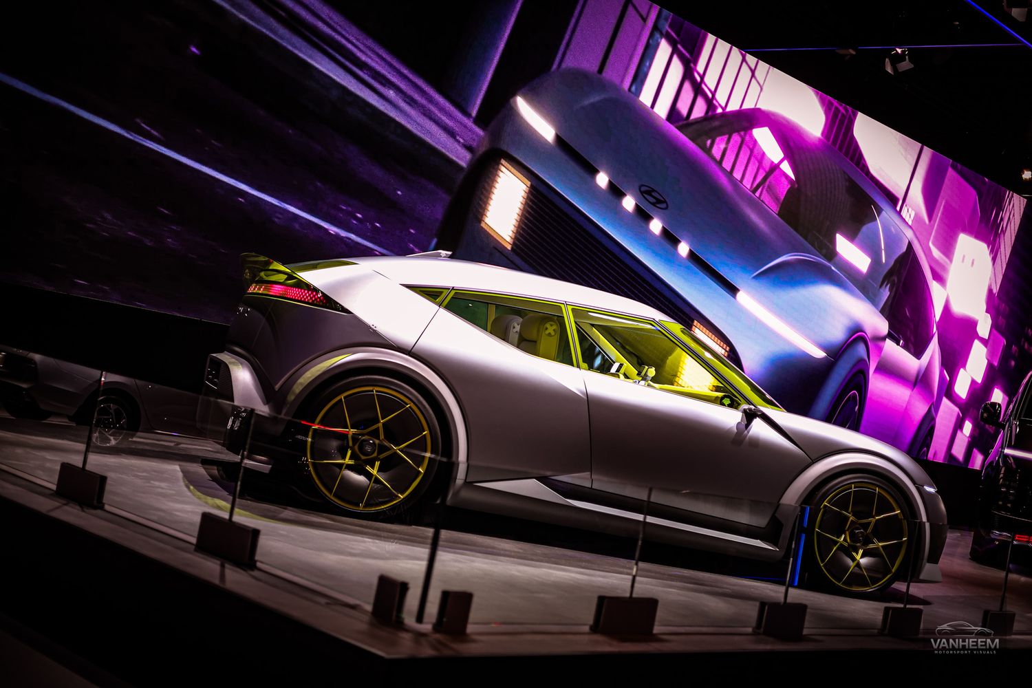 A sleek silver sports car displayed on an angled platform with dramatic purple and blue lighting in the background.