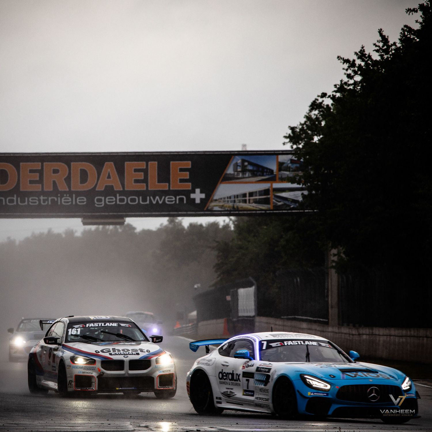 Two GT racing cars battle in wet conditions on a rain-soaked circuit beneath a Vanderdaele industrial buildings sponsor banner.