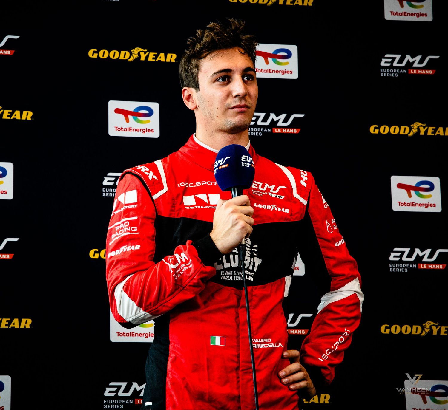 Racing driver in red suit holds microphone at ELMS European Le Mans Series event with Goodyear and TotalEnergies branding.