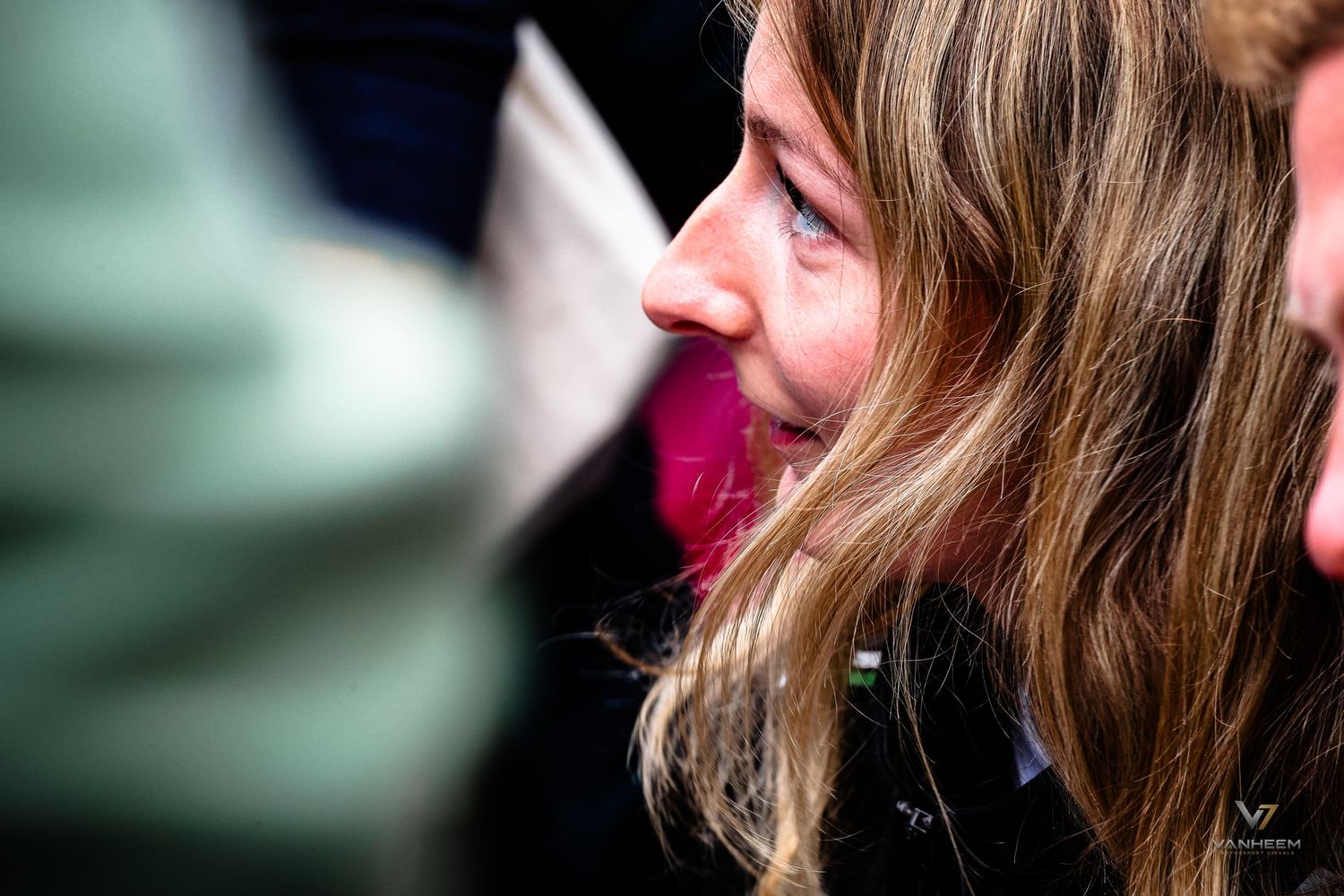 A young woman with long wavy hair smiles softly in a candid outdoor crowd photo.