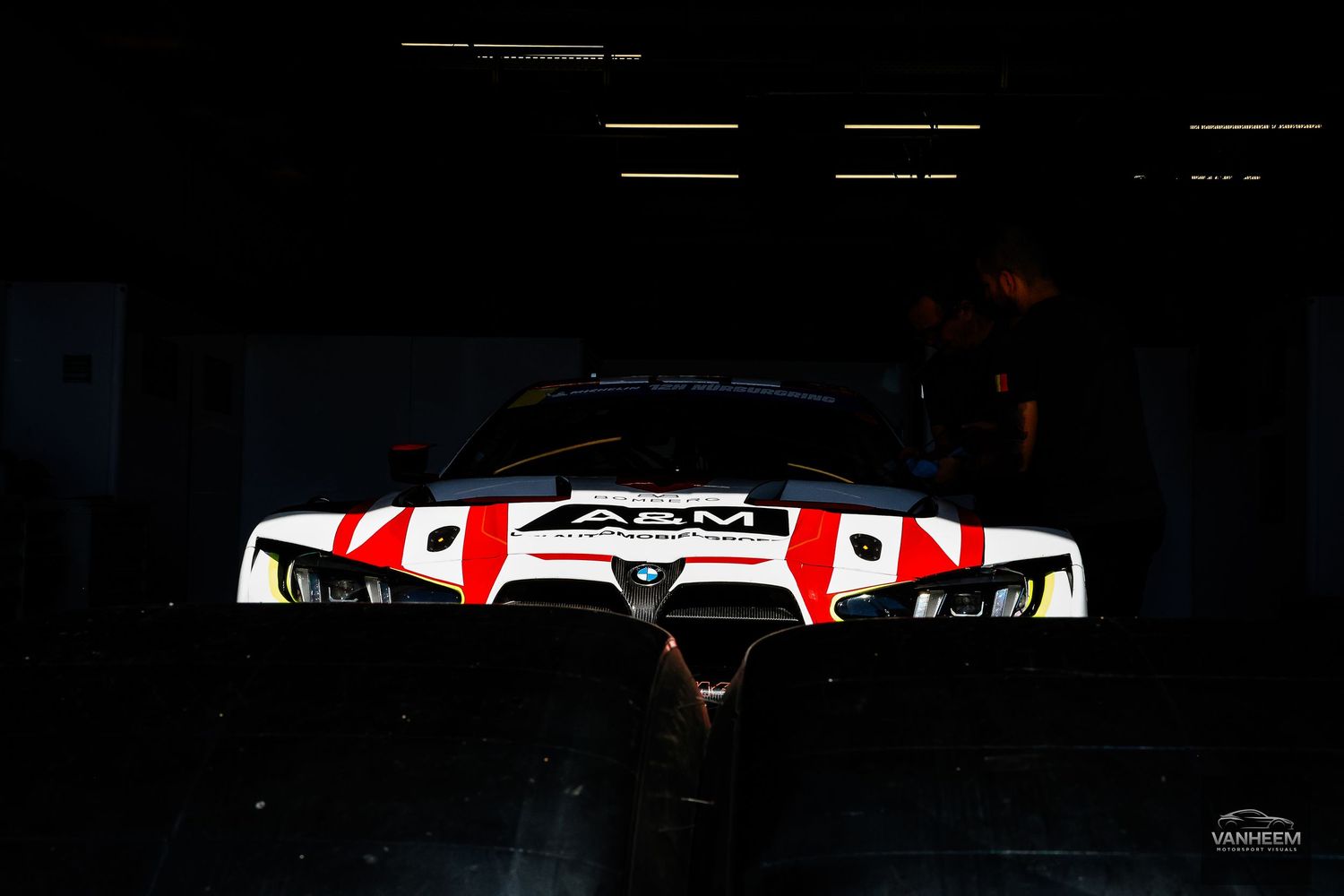 A&M racing car with red and white livery dramatically lit in a dark garage pit lane.
