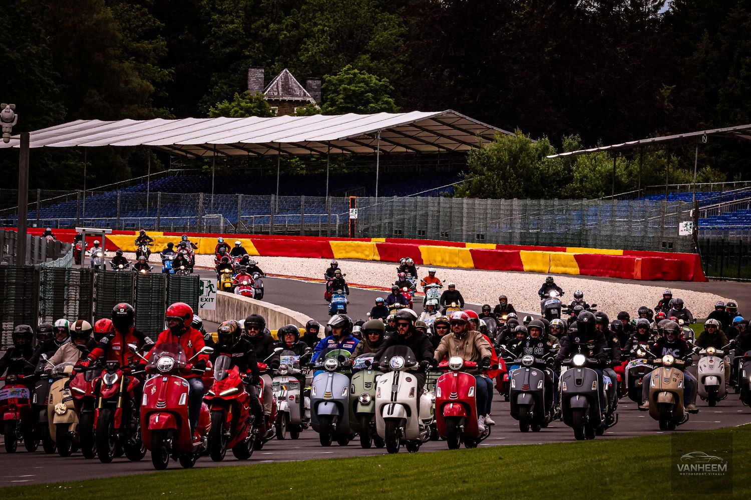 Large group of motorcycles and scooters lined up on a racing circuit surrounded by lush green trees.