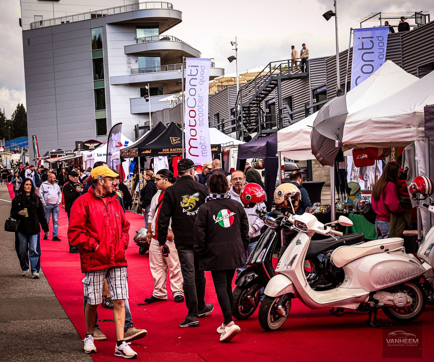 Crowd gathered on red carpet at outdoor motorcycle expo with scooters on display and vendor tents lining the walkway.