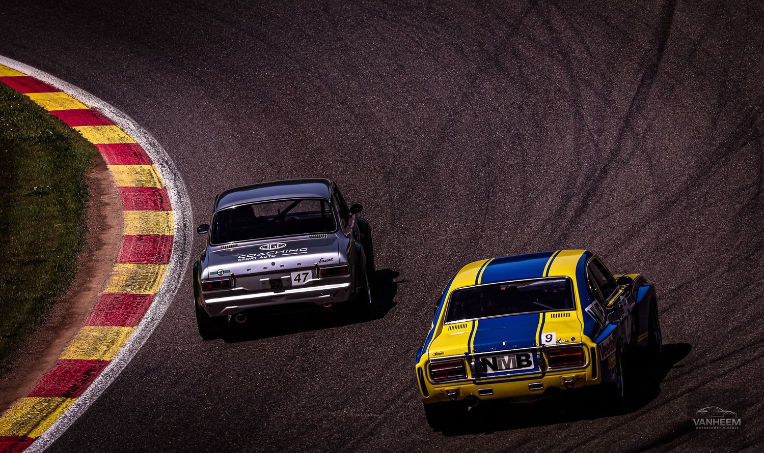Two vintage race cars, yellow-blue and dark, compete on a winding asphalt track during a classic car racing event.