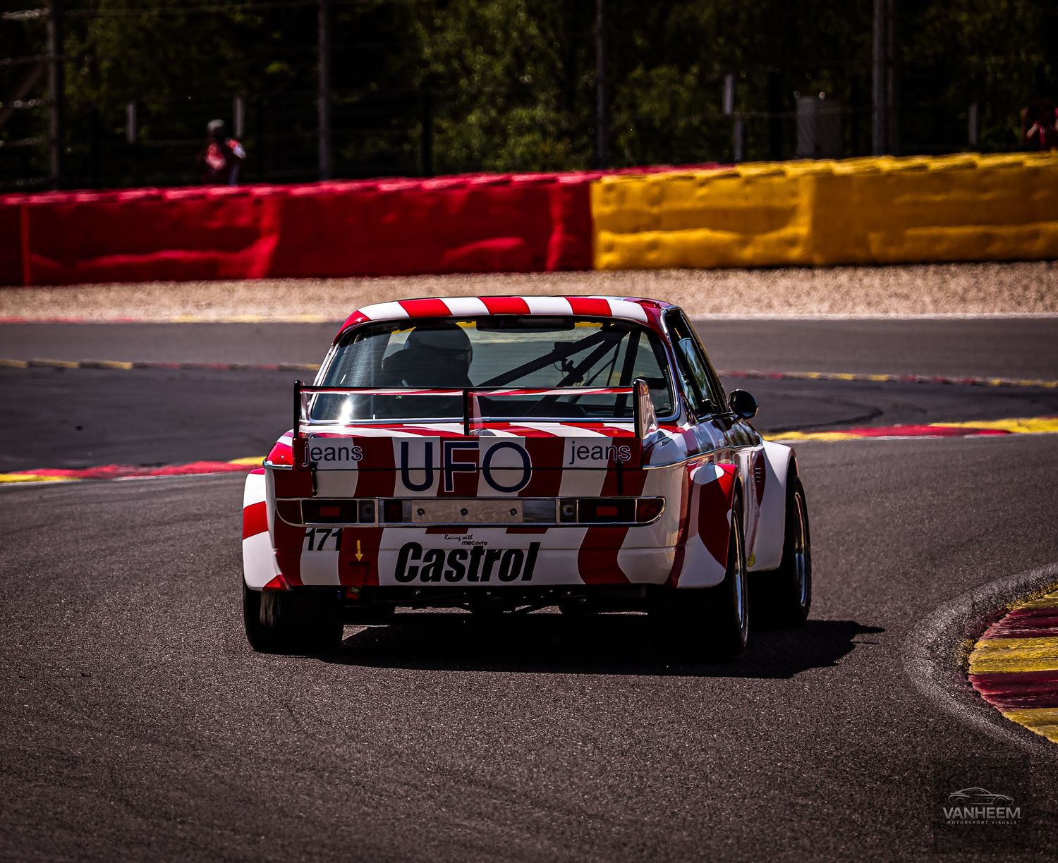 A vintage race car with Castrol and UFO livery competing on a circuit track with colorful barriers in the background.