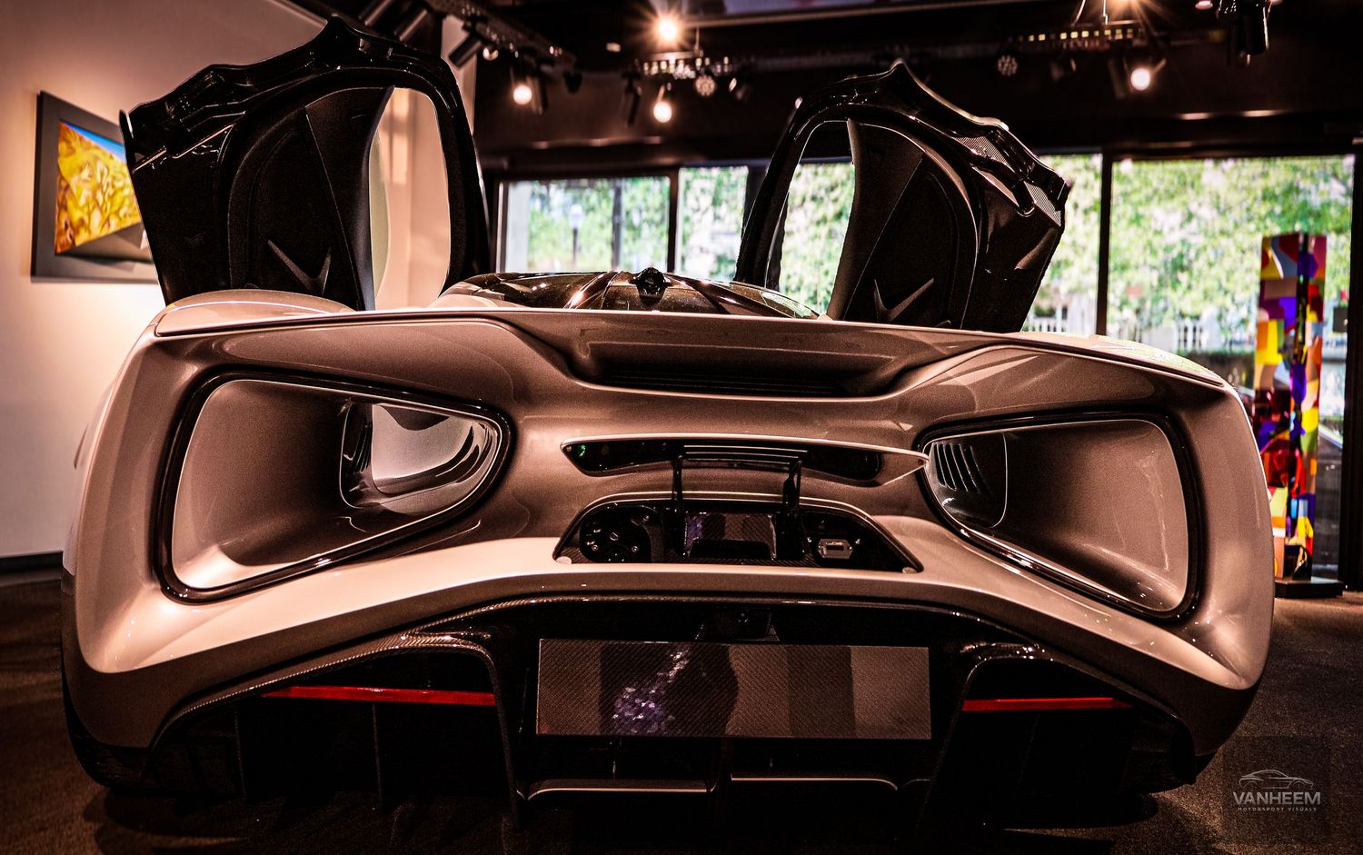 Rear view of a sleek futuristic supercar with open gull-wing doors displayed in a modern art gallery showroom.