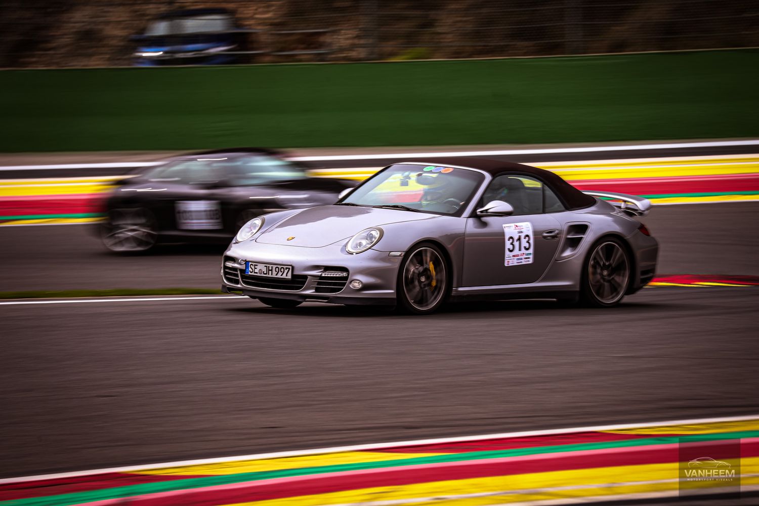 A silver Porsche 911 Turbo convertible racing on the Spa-Francorchamps circuit with a blurred car in the background.