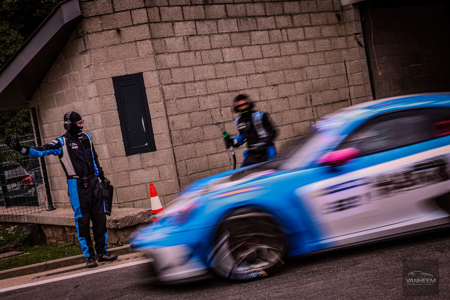 Two marshals stand roadside as a blue rally car speeds past a concrete wall during a motorsport event.