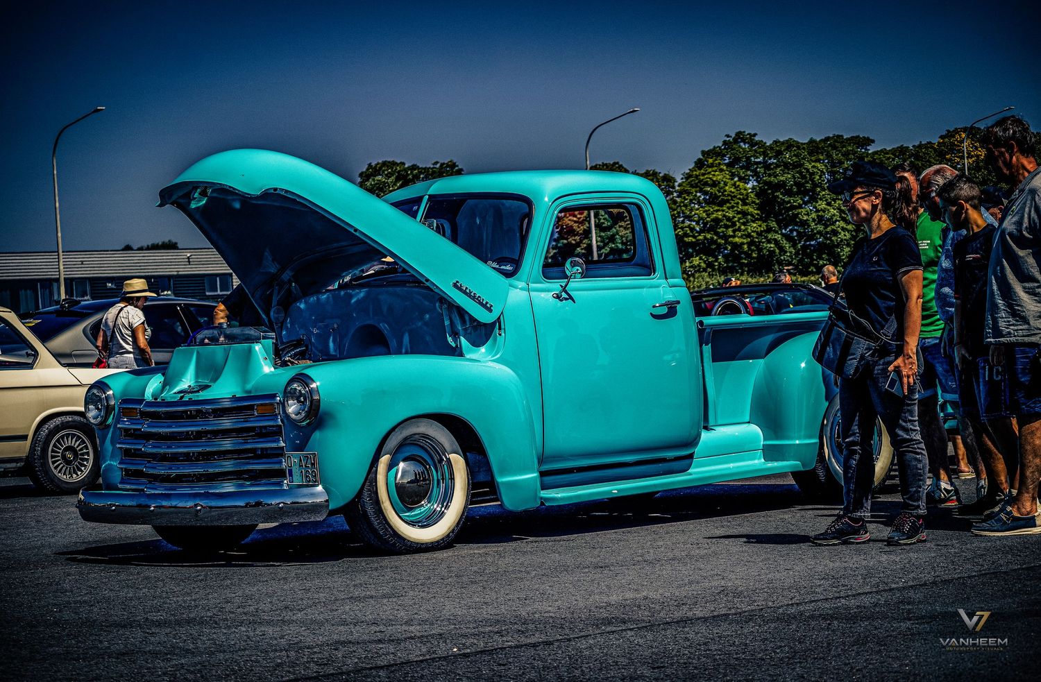Vintage teal lowrider pickup truck with hood open displayed at outdoor car show with admirers gathered around.