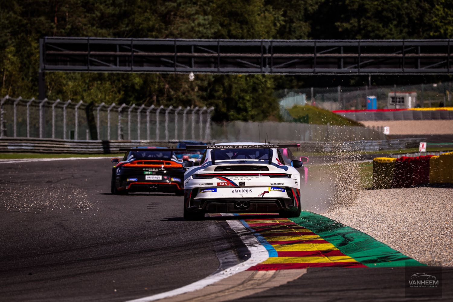 Race cars navigate a challenging turn on a professional racing circuit with colorful track markings and gravel runoff areas.