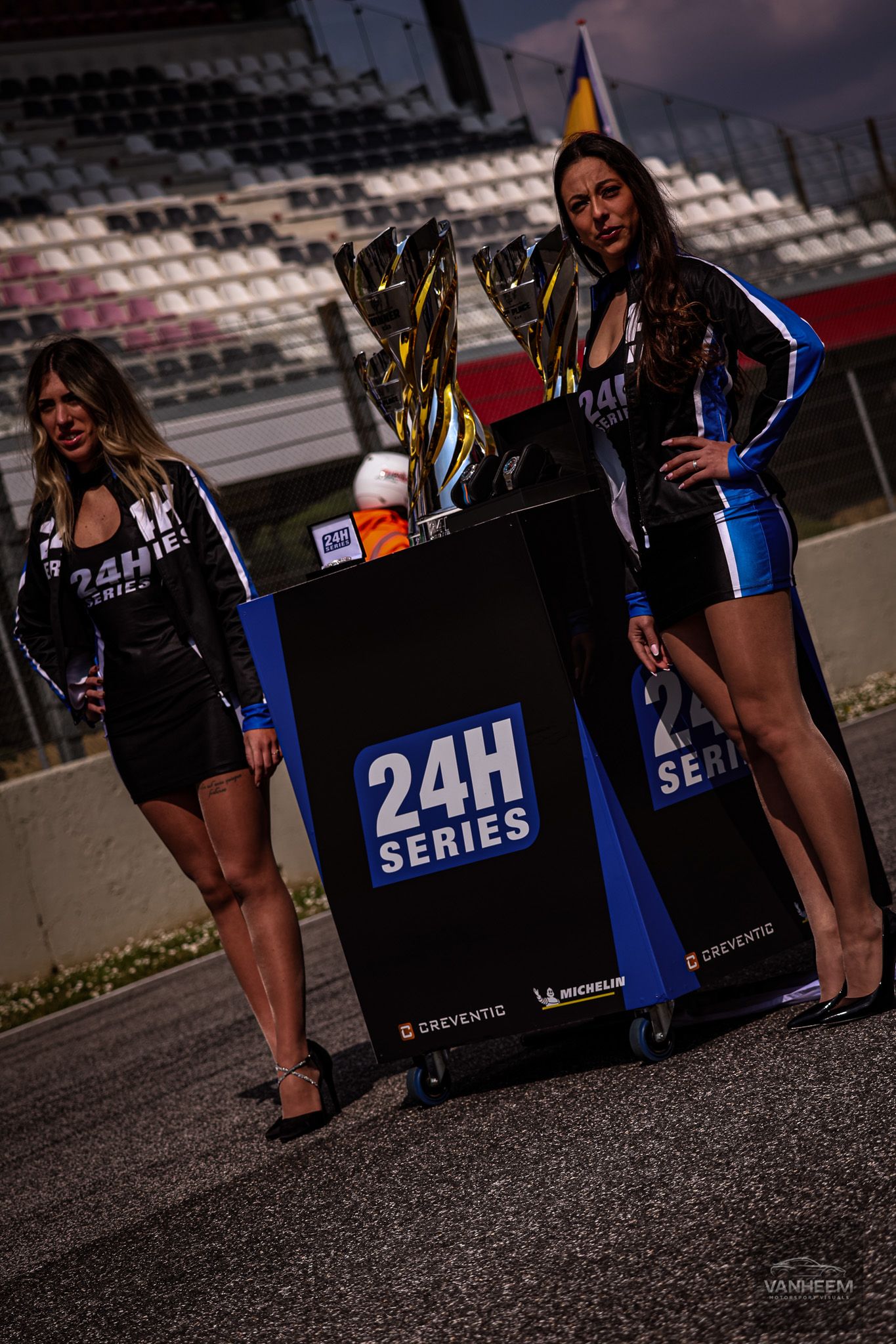 Grid girls pose with Series 24h racing signage at a motorsport event during evening hours.
