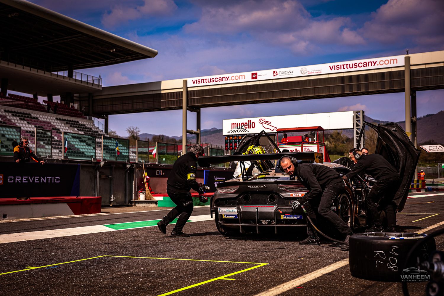 Racing pit crew performs maintenance on a Formula 1 car during a pit stop at dusk under dramatic sky.