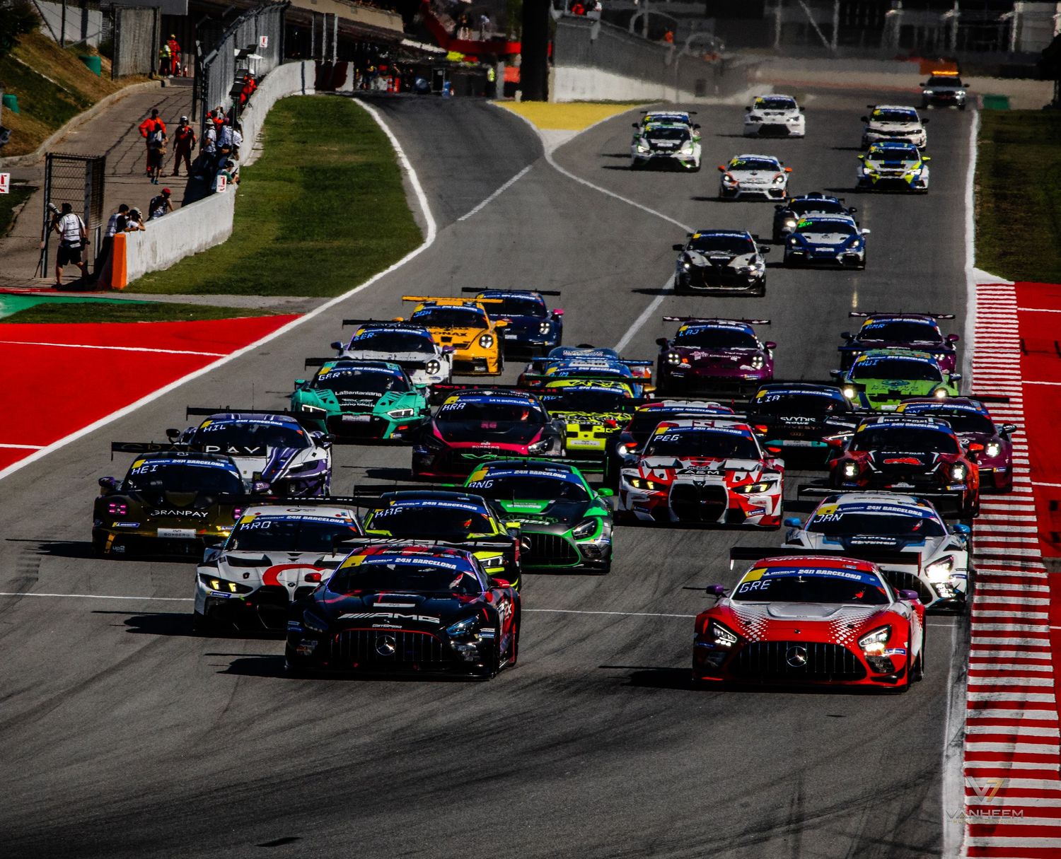 A large field of GT racing cars navigate a sweeping corner at the start of a motorsport race on a scenic circuit.