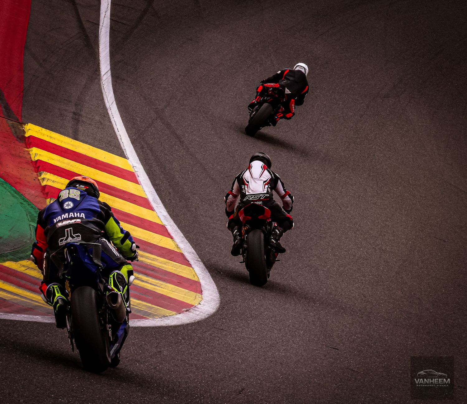 Three MotoGP riders race on a colorful circuit track, with Yamaha leading two red motorcycles through a curve.