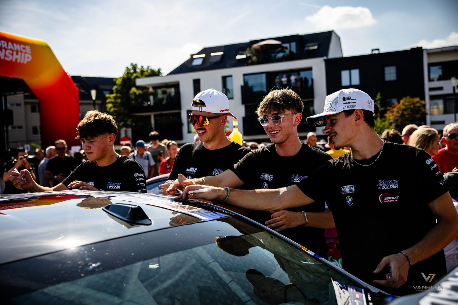 Four young racing drivers sign a car hood at an outdoor motorsport event with crowds gathered in the background.
