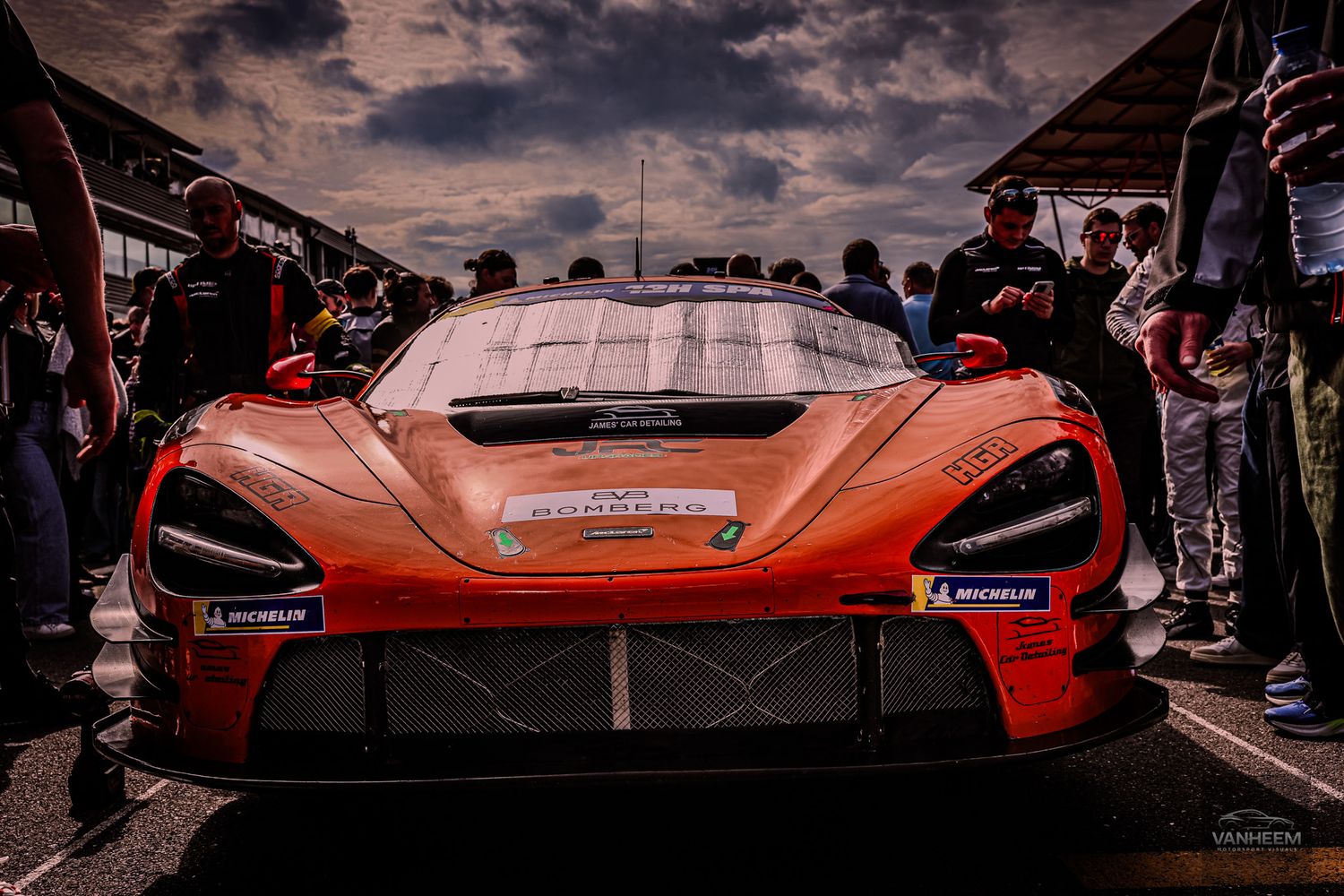 Orange McLaren GT3 race car on pit lane surrounded by crew members under dramatic cloudy skies at motorsport event.