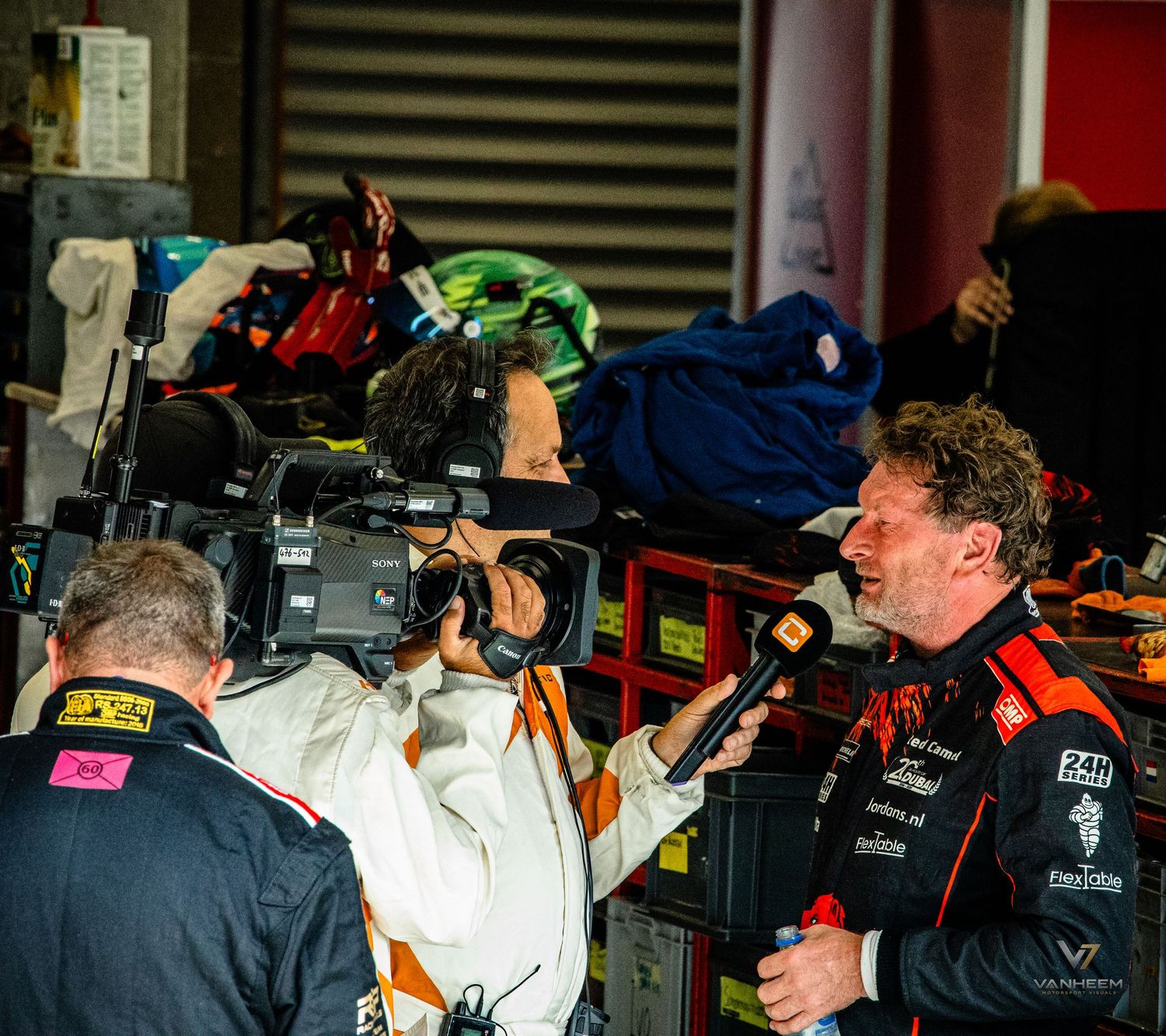 Racing team member being interviewed by TV crew with camera in busy pit lane during motorsport event.