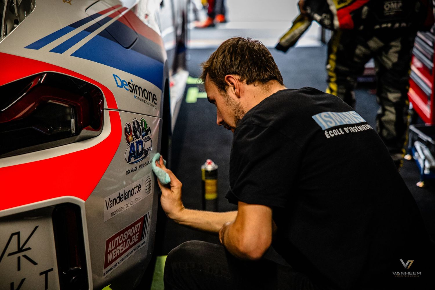 Mechanic in black shirt polishing a racing car livery in a pit garage during a motorsport event.