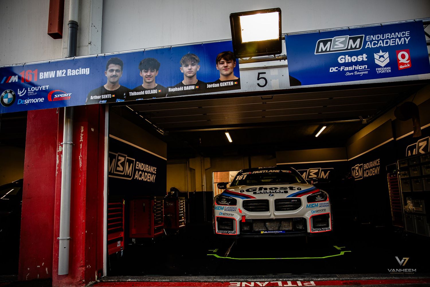 BMW M2 racing car in pit garage at endurance race, team members pictured above on banner, race setup visible.