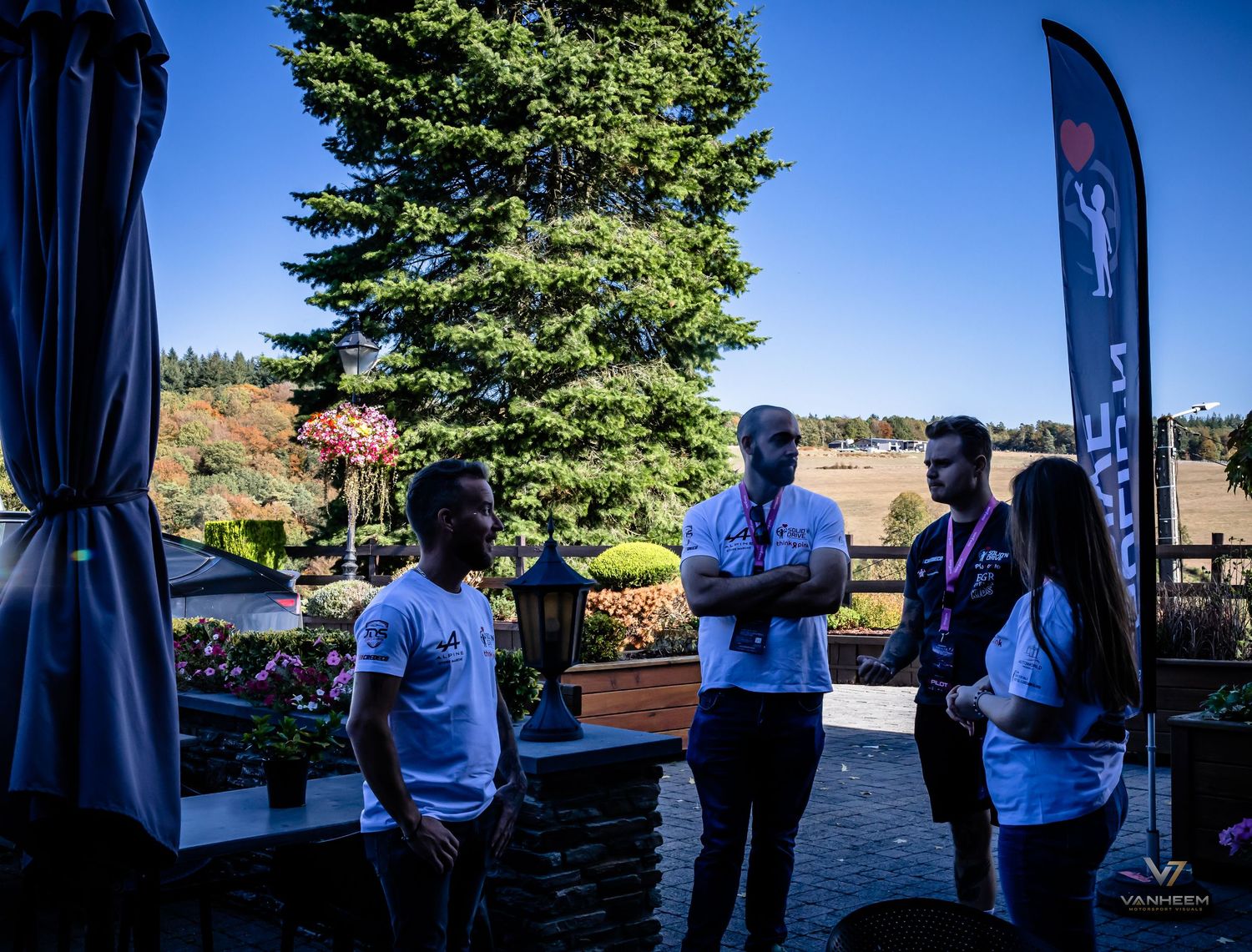 Group of people conversing outdoors near a decorative banner and tall evergreen tree at a sunny event venue.