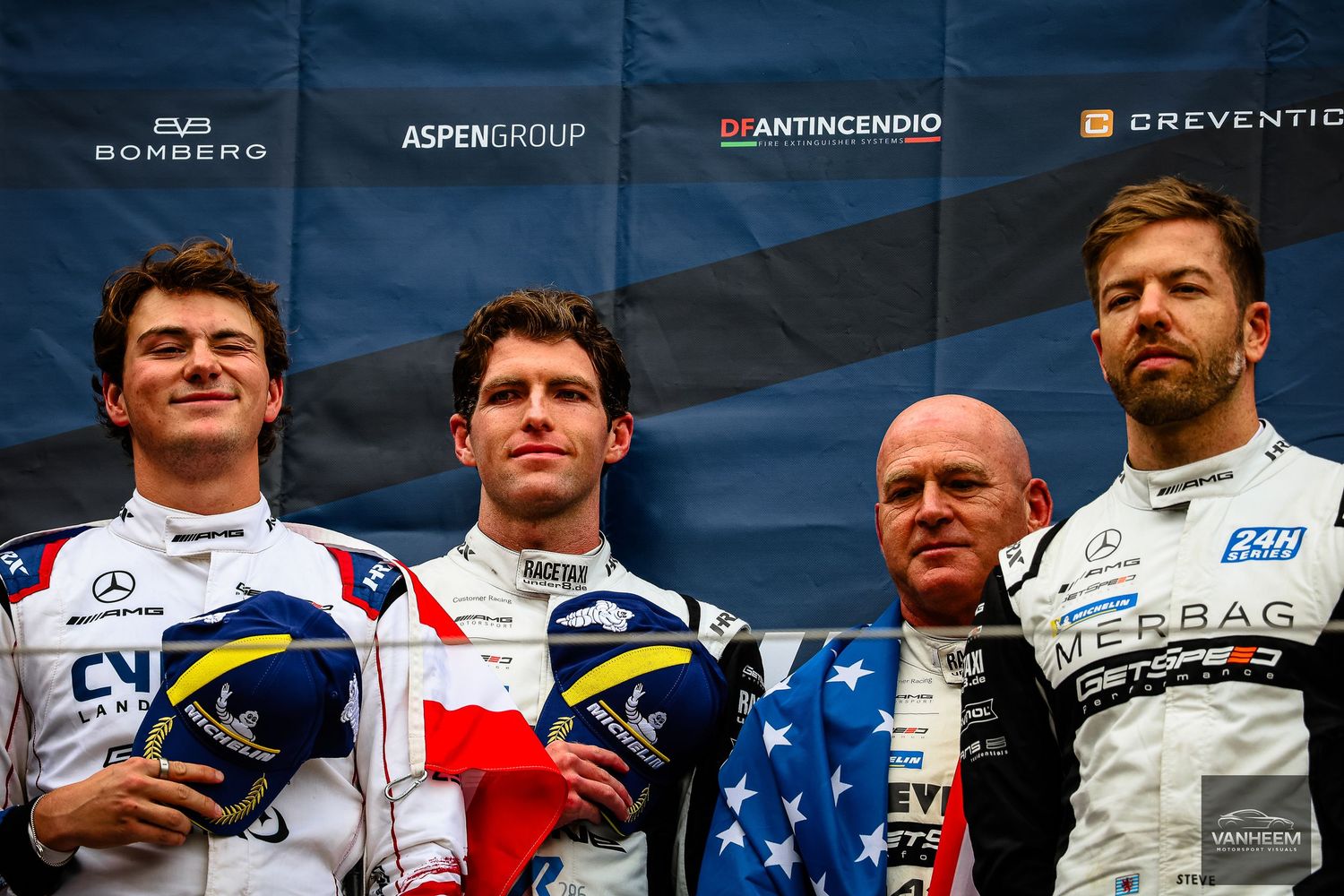 Four racing drivers pose on podium with sponsor banners including Aspen Group and Bomberg at motorsport event.