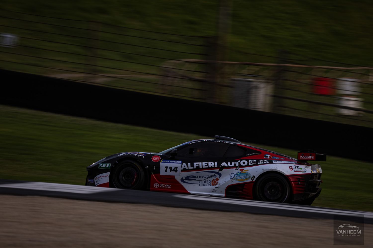 Race car speeds along dark racetrack with red and white livery during competitive motorsport event.