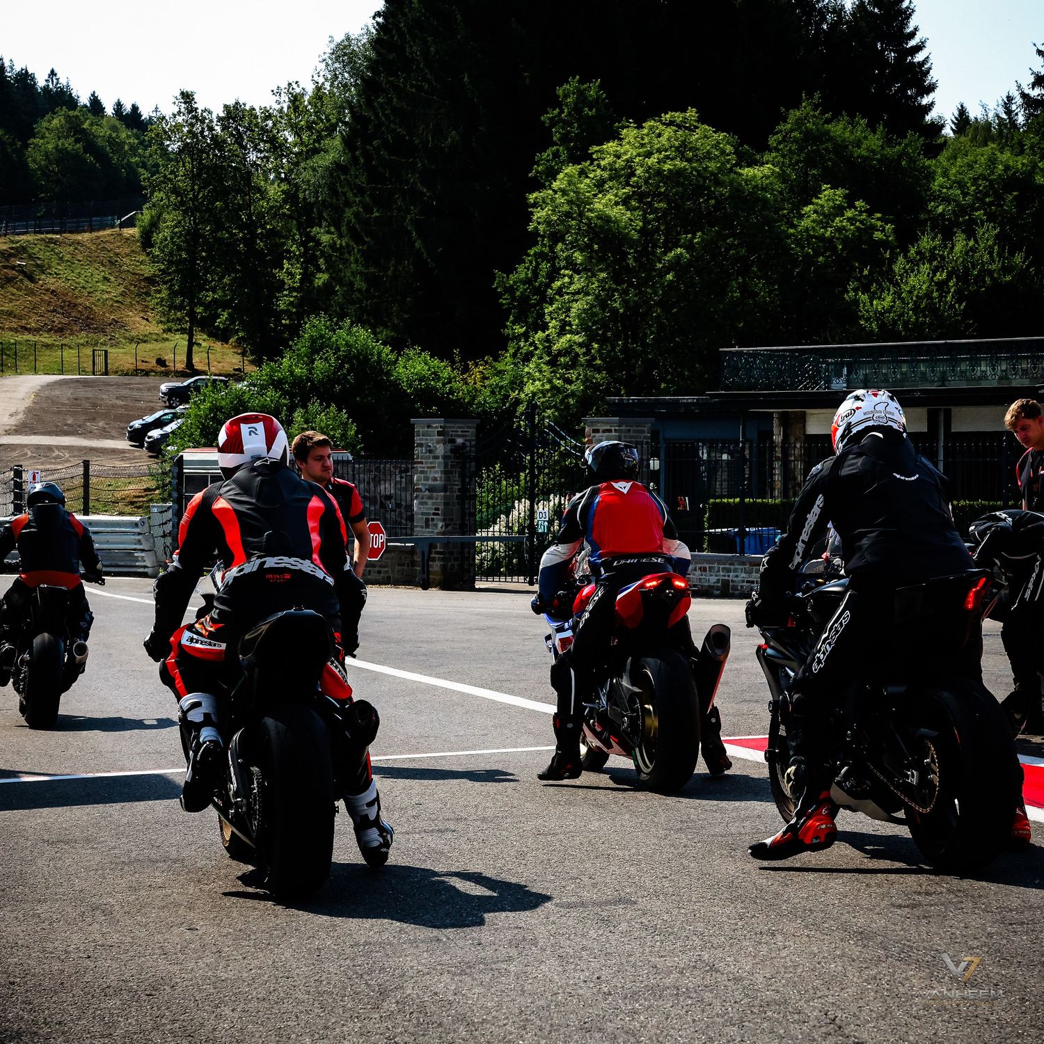 Motorcyclists in racing gear gather on a track circuit surrounded by lush green trees on a sunny day.