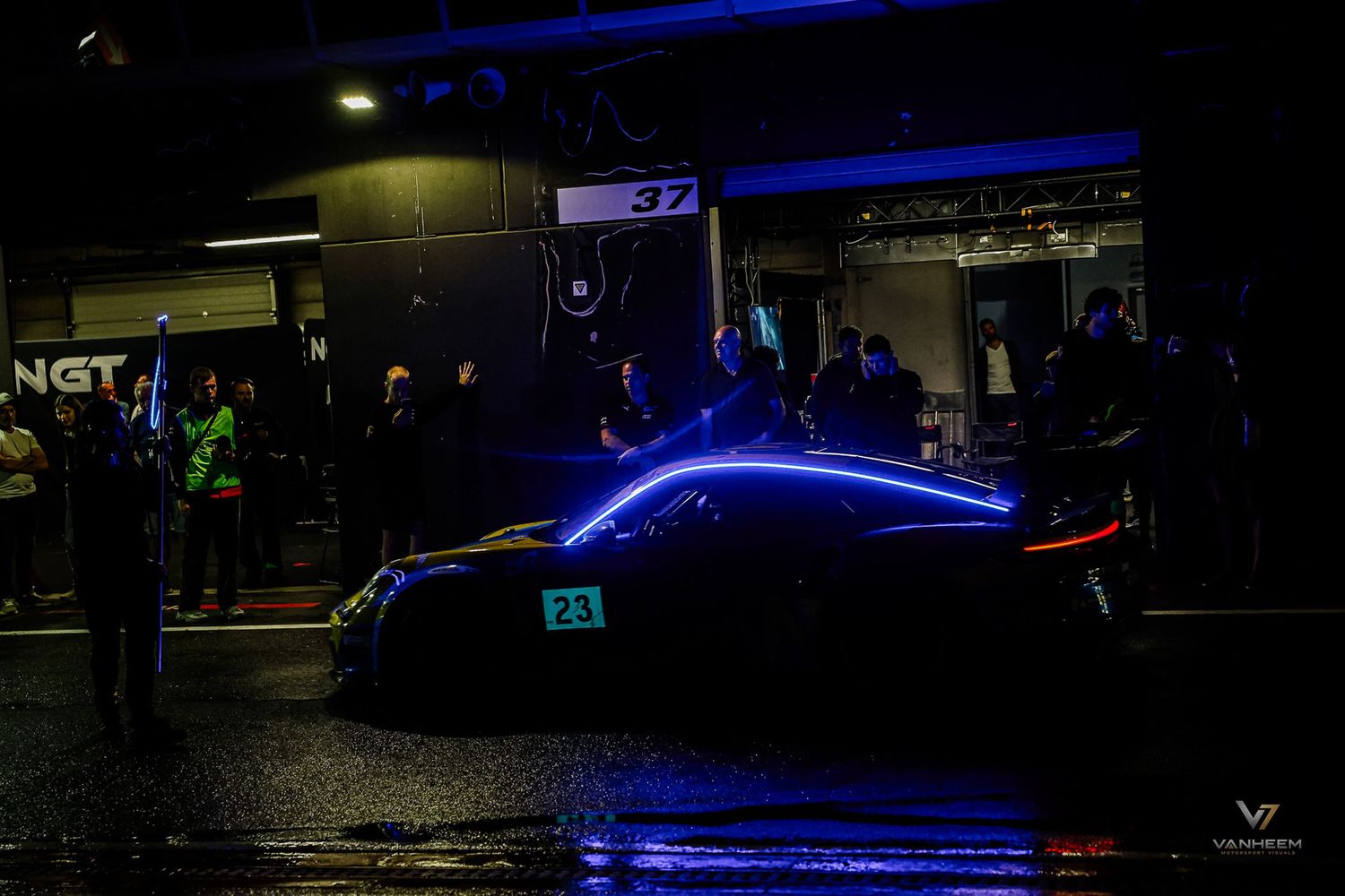 A glowing race car with blue LED lights sits in pit lane at night during a motorsport endurance racing event.