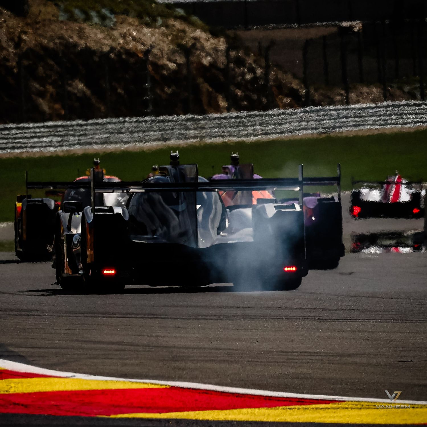Racing cars compete on a circuit track, with smoke trailing from the lead black LMP prototype race car during an endurance race.