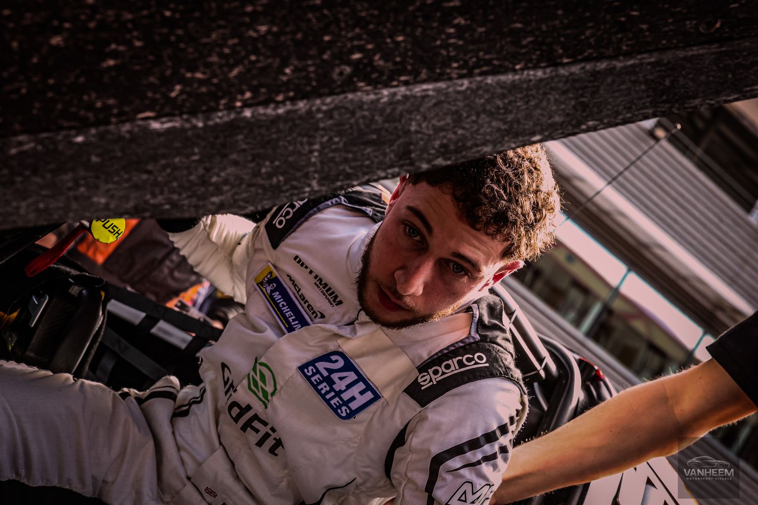A young male racing driver in a white Sparco suit with 24H Series patches looks up from his race car cockpit.
