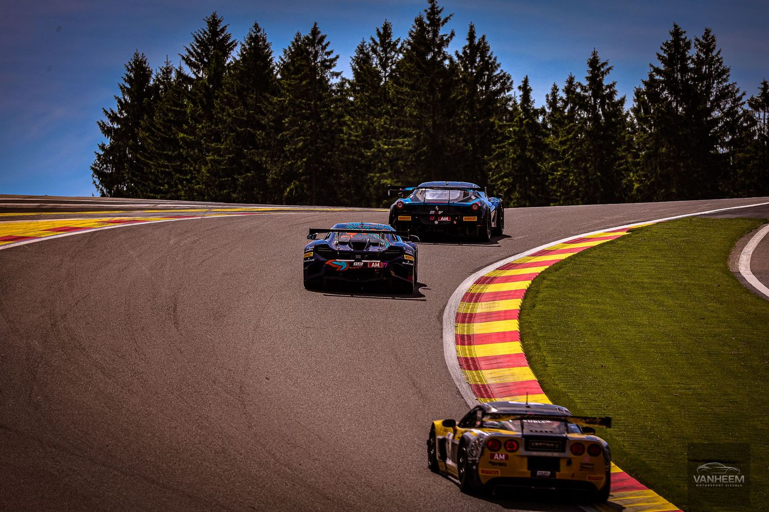 Three racing cars navigate a curved track at Spa-Francorchamps circuit, surrounded by tall pine trees under a blue sky.