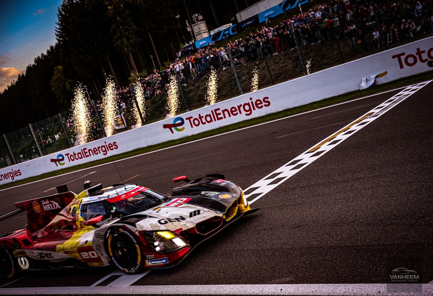 A Ferrari LMH race car speeds past the TotalEnergies banner at dusk with sparks flying at a WEC endurance race.