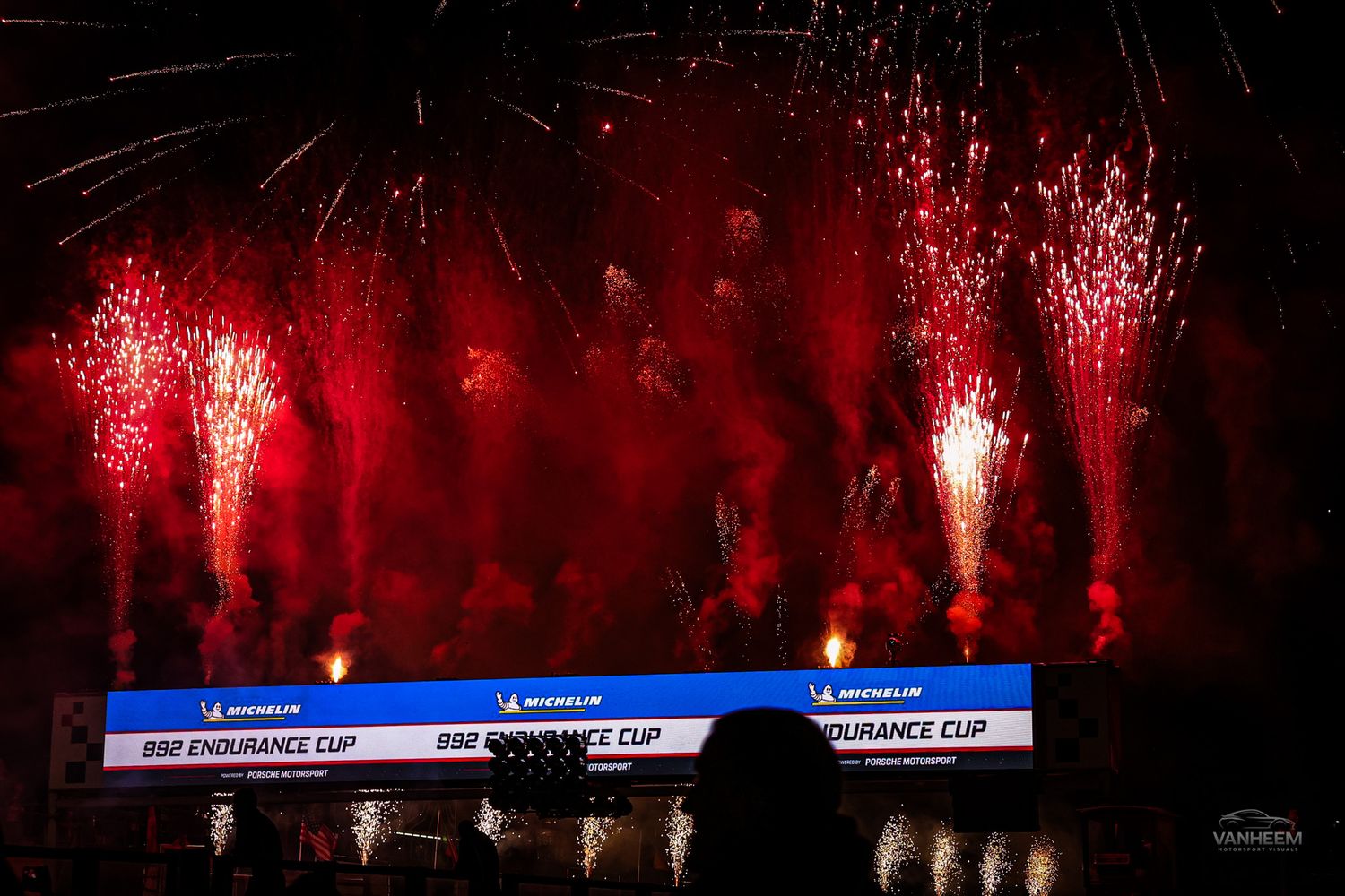 Spectacular red fireworks display illuminates the night sky above a racing circuit.