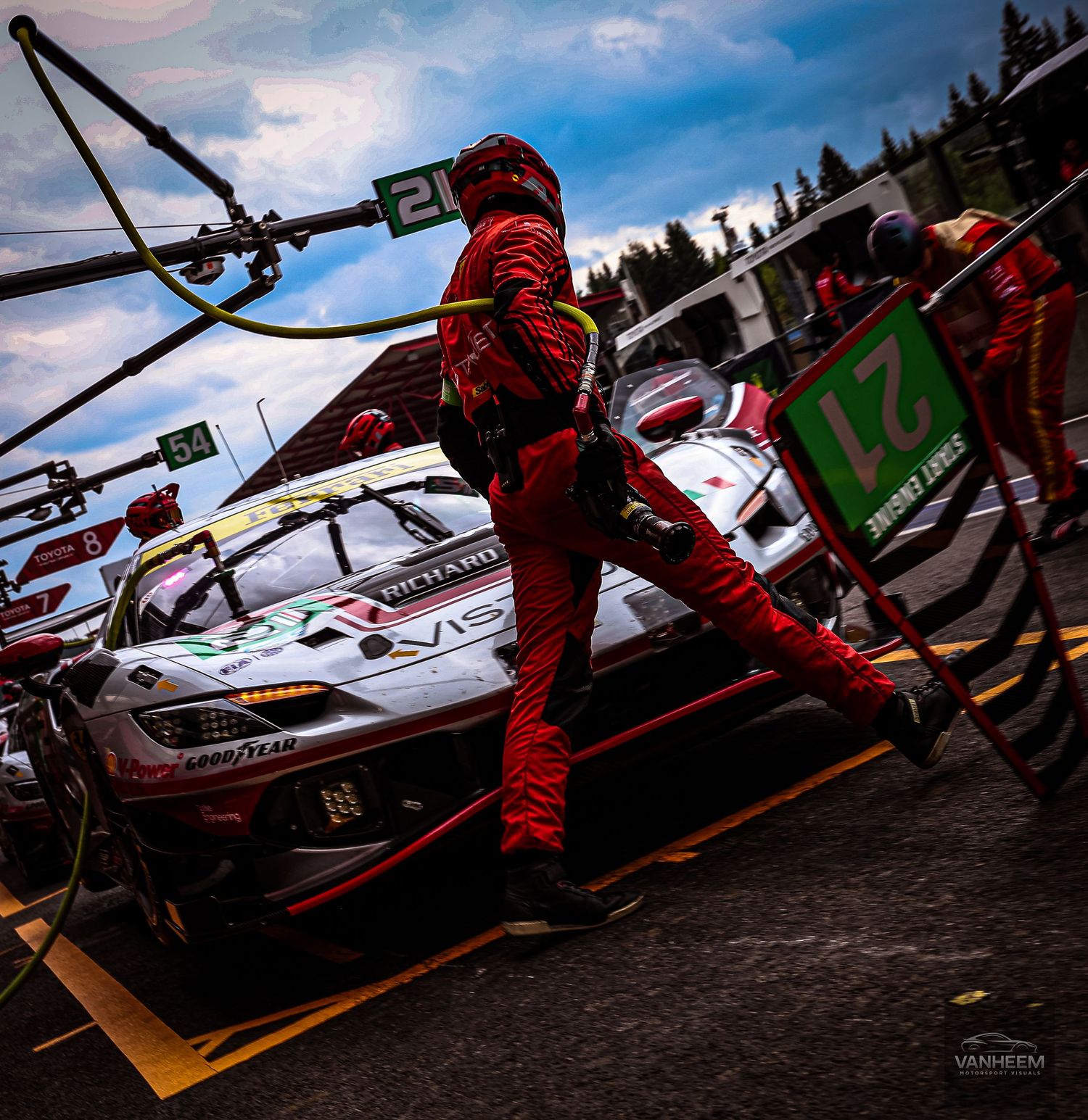 Red-suited pit crew member services a racing car during a motorsport event at a professional circuit under dramatic cloudy skies.