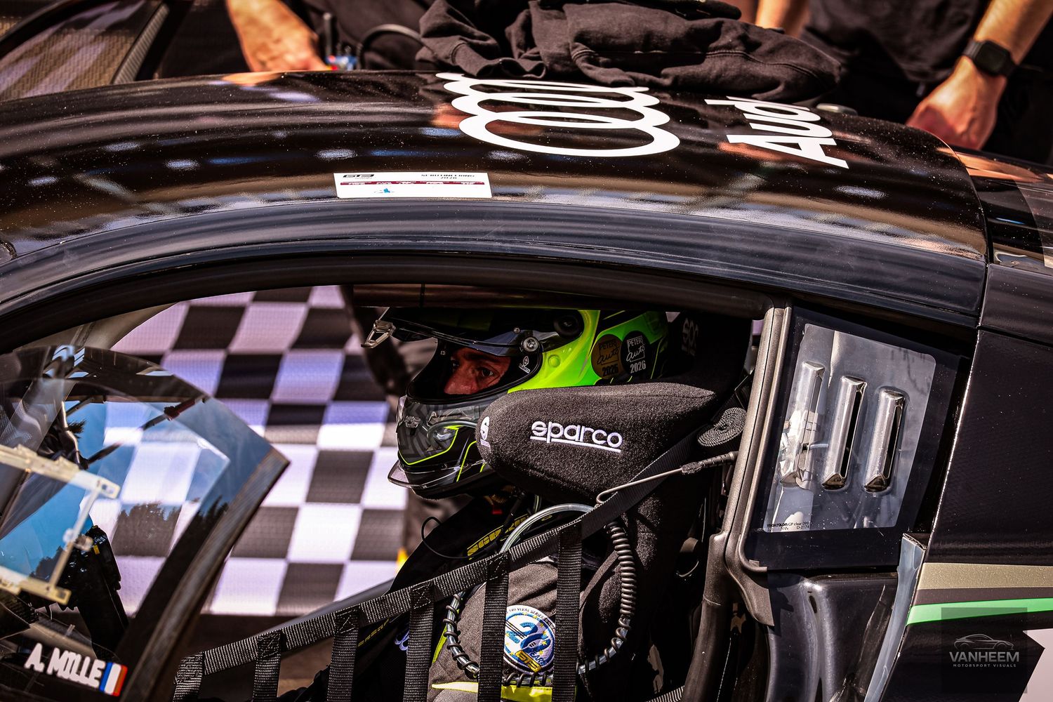 A racing driver in a Sparco helmet sits inside a black race car cockpit with checkered flag pattern visible.