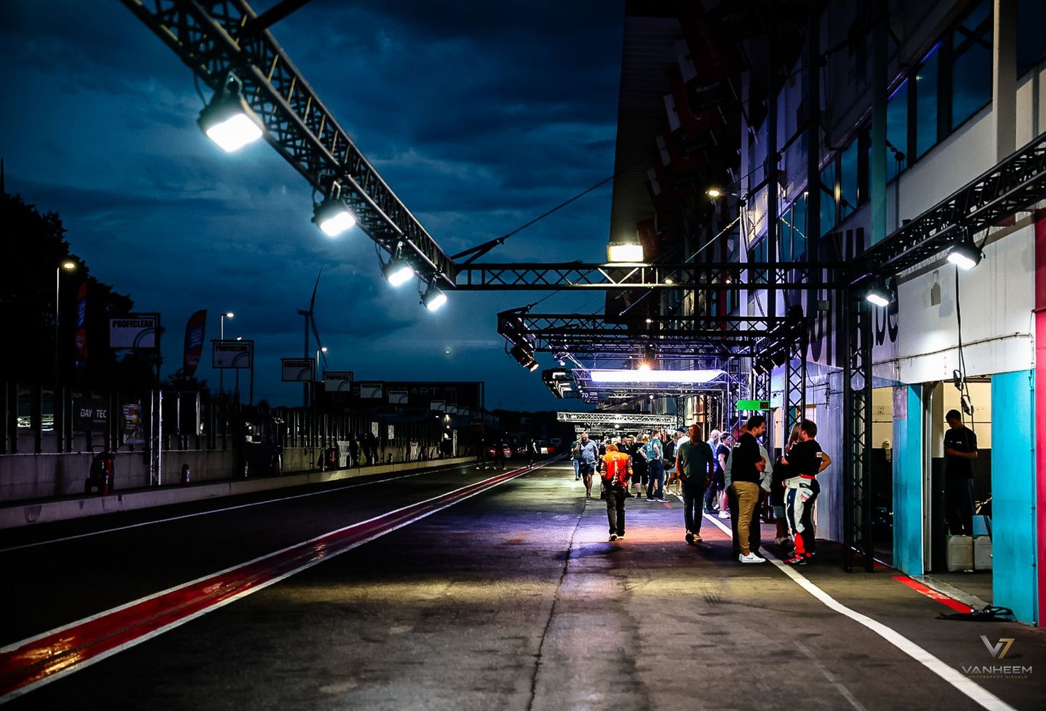Night scene at a motorsport pit lane with crew members walking under bright lights during a racing event.