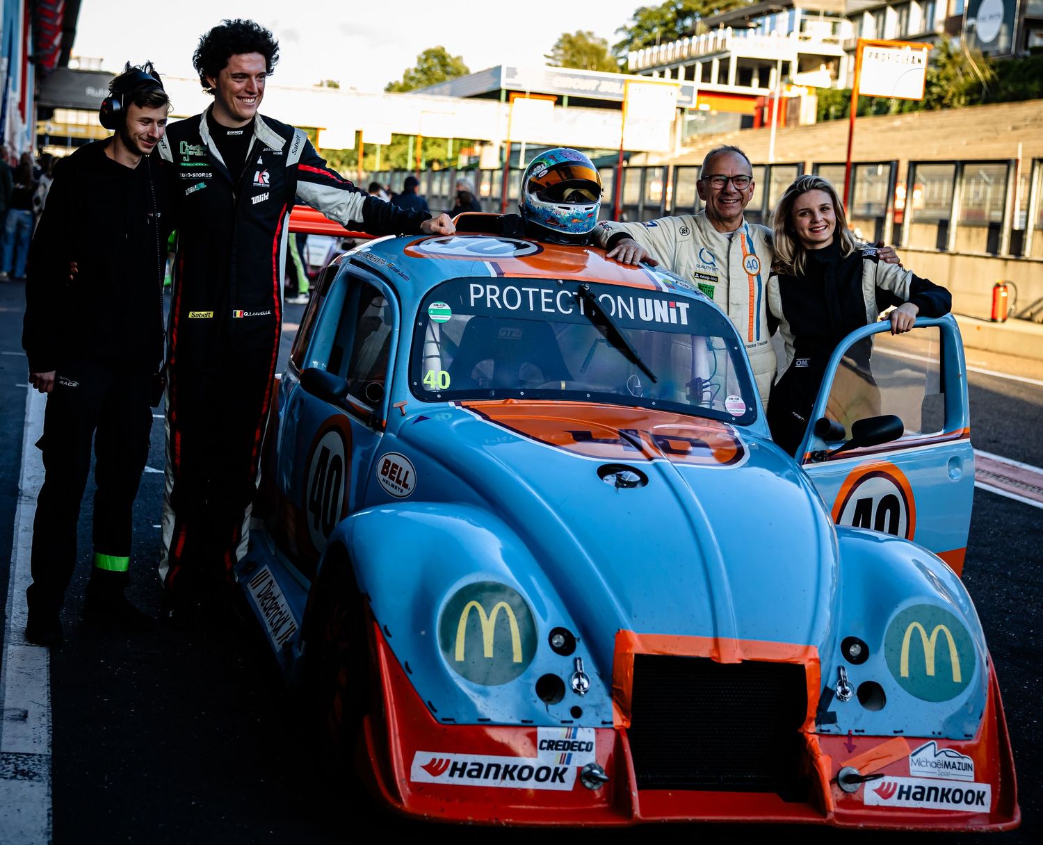 Four people posing with a Gulf-liveried VW Beetle race car featuring McDonald's sponsorship on a racing circuit pit lane.